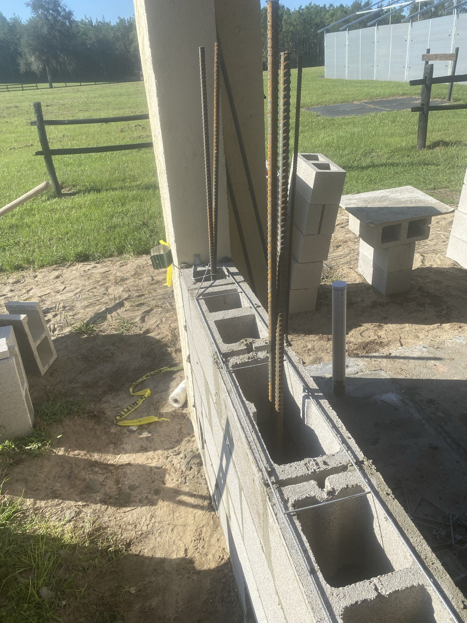 A partially laid concrete block stem wall with vertical rebar and fresh mortar joints runs beside a stucco column, with green pasture and board fencing beyond
