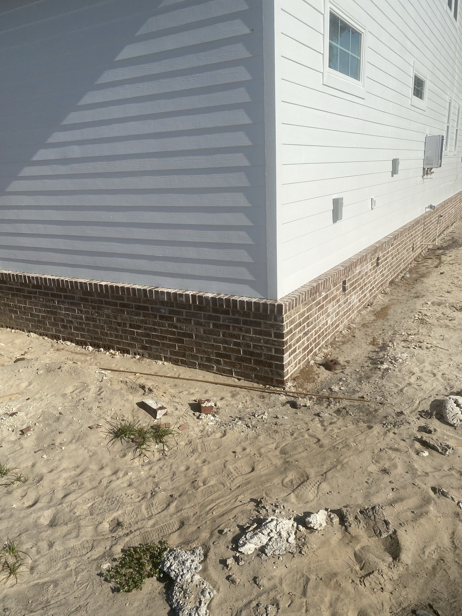 Close view of the corner of a house where a brick foundation wall with a soldier-course cap meets white lap siding, over sandy ground