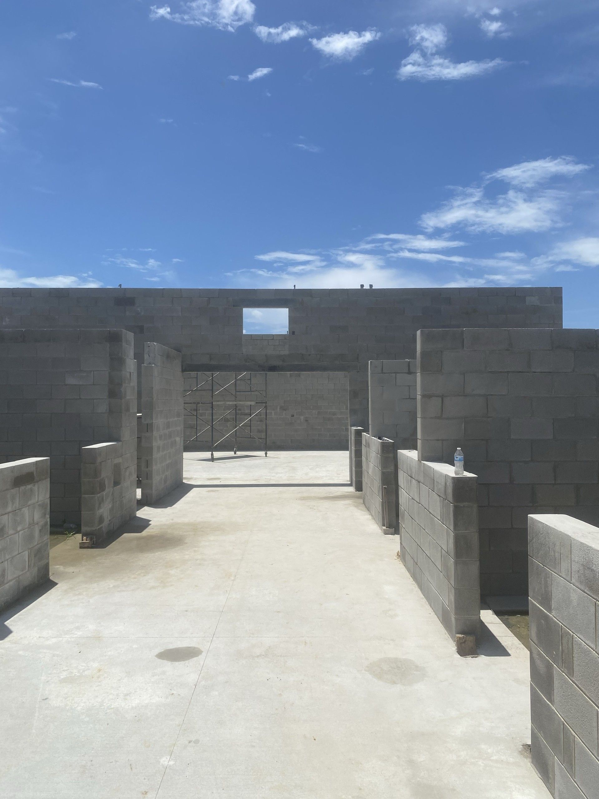 A corridor between freshly laid concrete block interior walls of a roofless building shell, with open blue sky above and rebar visible at the wall tops