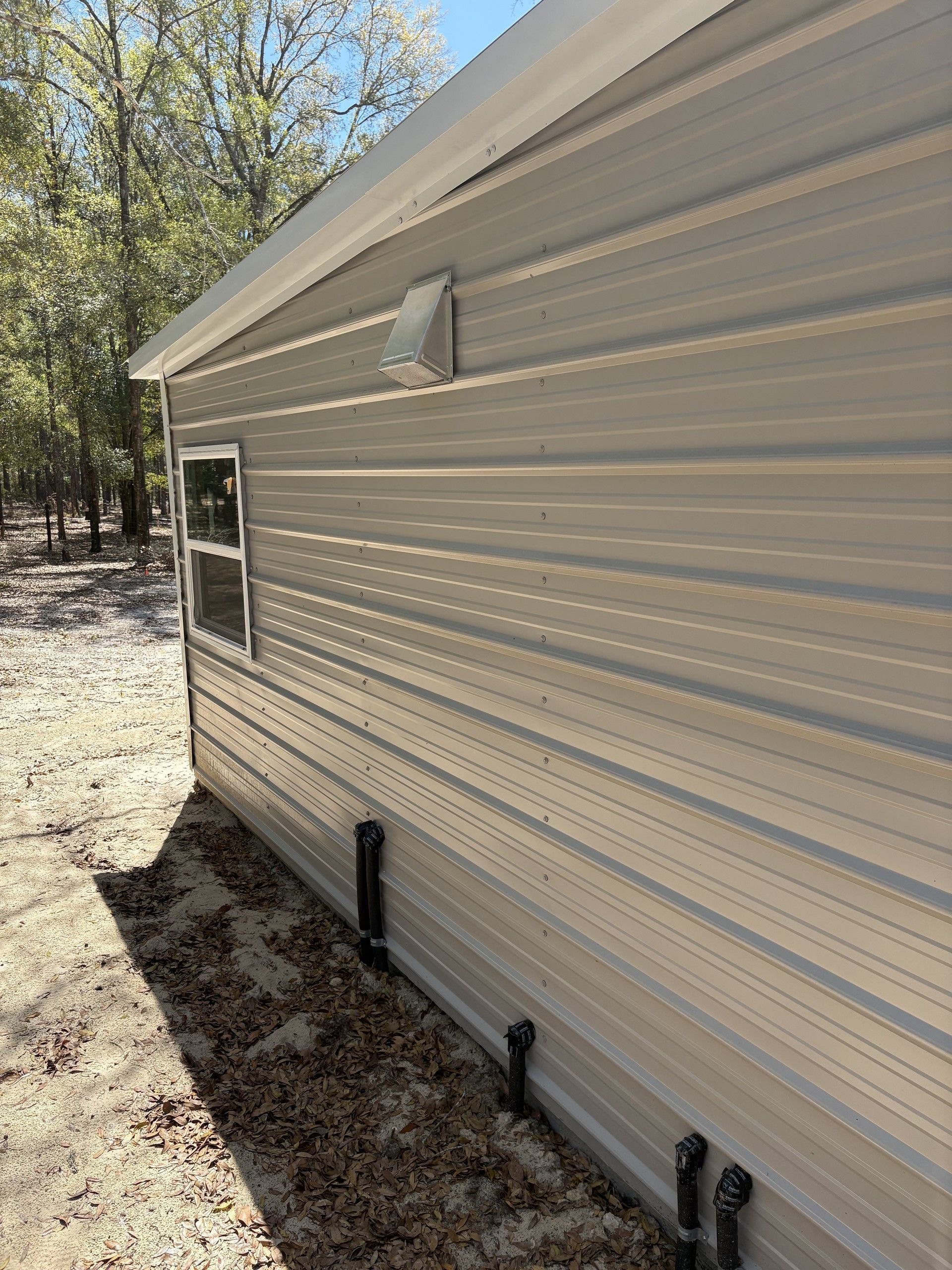 The side wall of a completed tan metal building with a metal roof overhang, a dryer vent hood, a single window, and black drainage stub-outs along the base, set on sandy ground among pine and oak trees