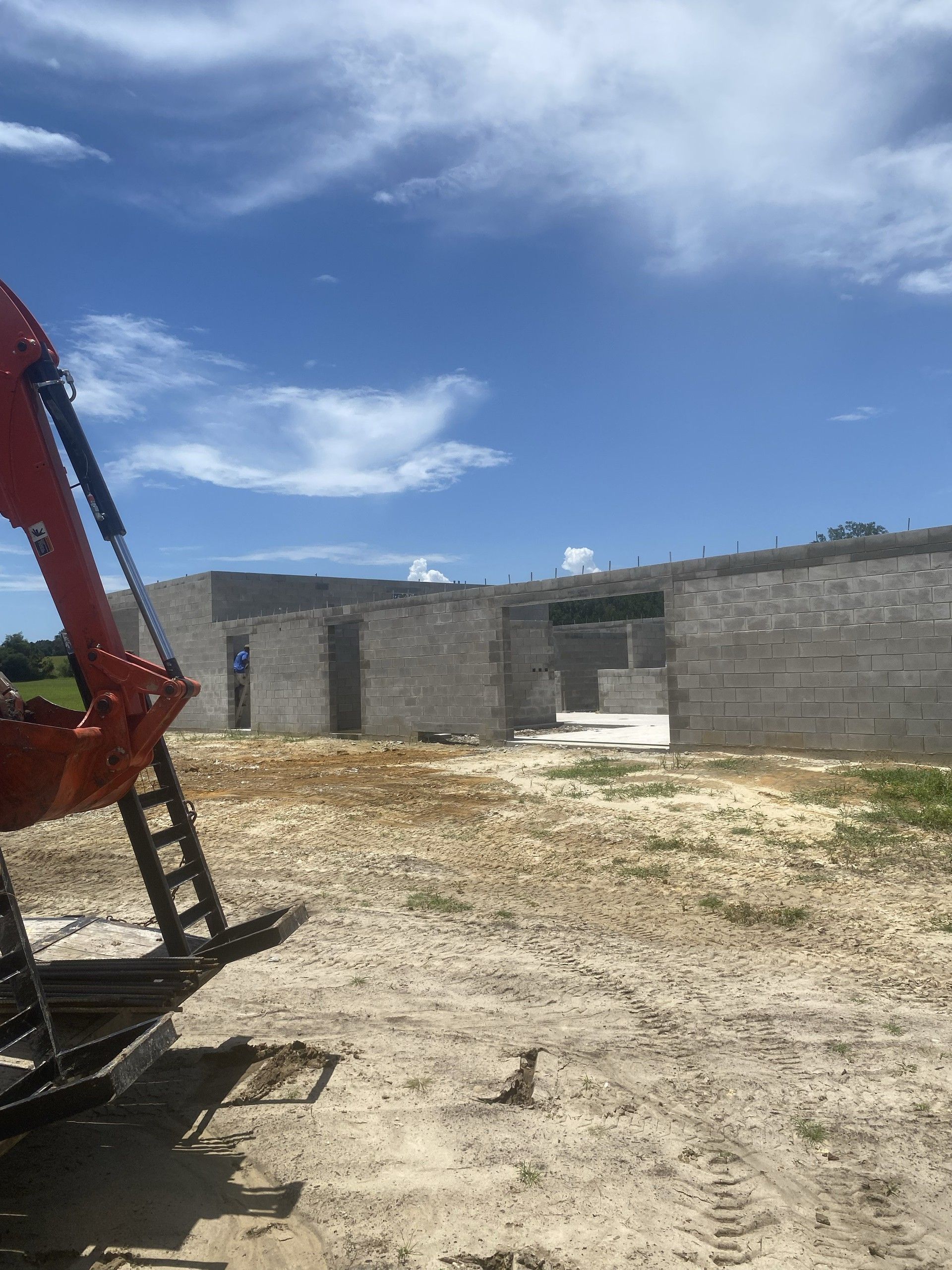 A yellow excavator arm crosses the foreground of a site where tall concrete block walls of a long single-story building stand under a blue sky, with a worker in a doorway