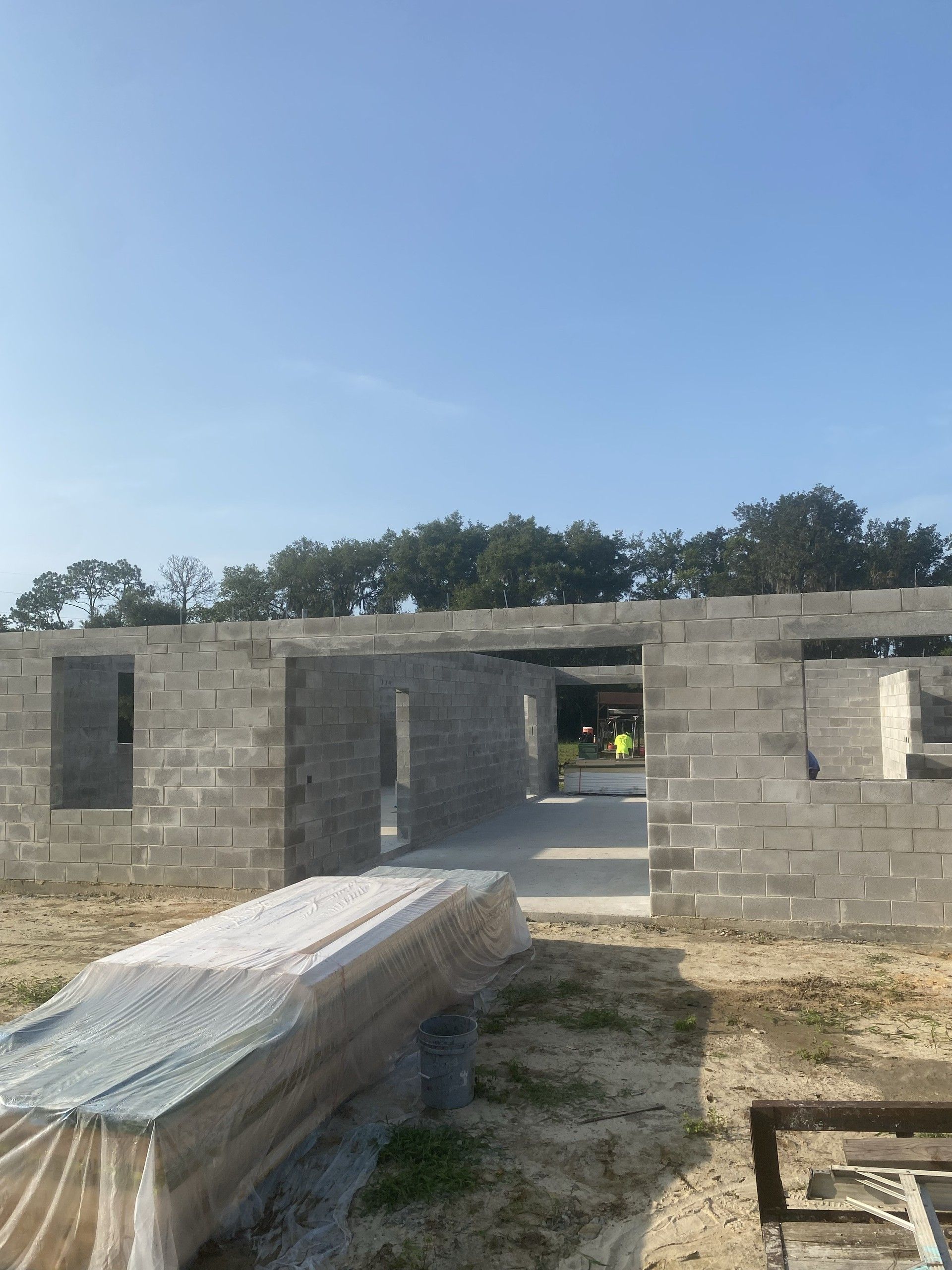 The front wall of a concrete block building shell with several window and door openings, a plastic-covered bundle of lumber stacked on the sand in front
