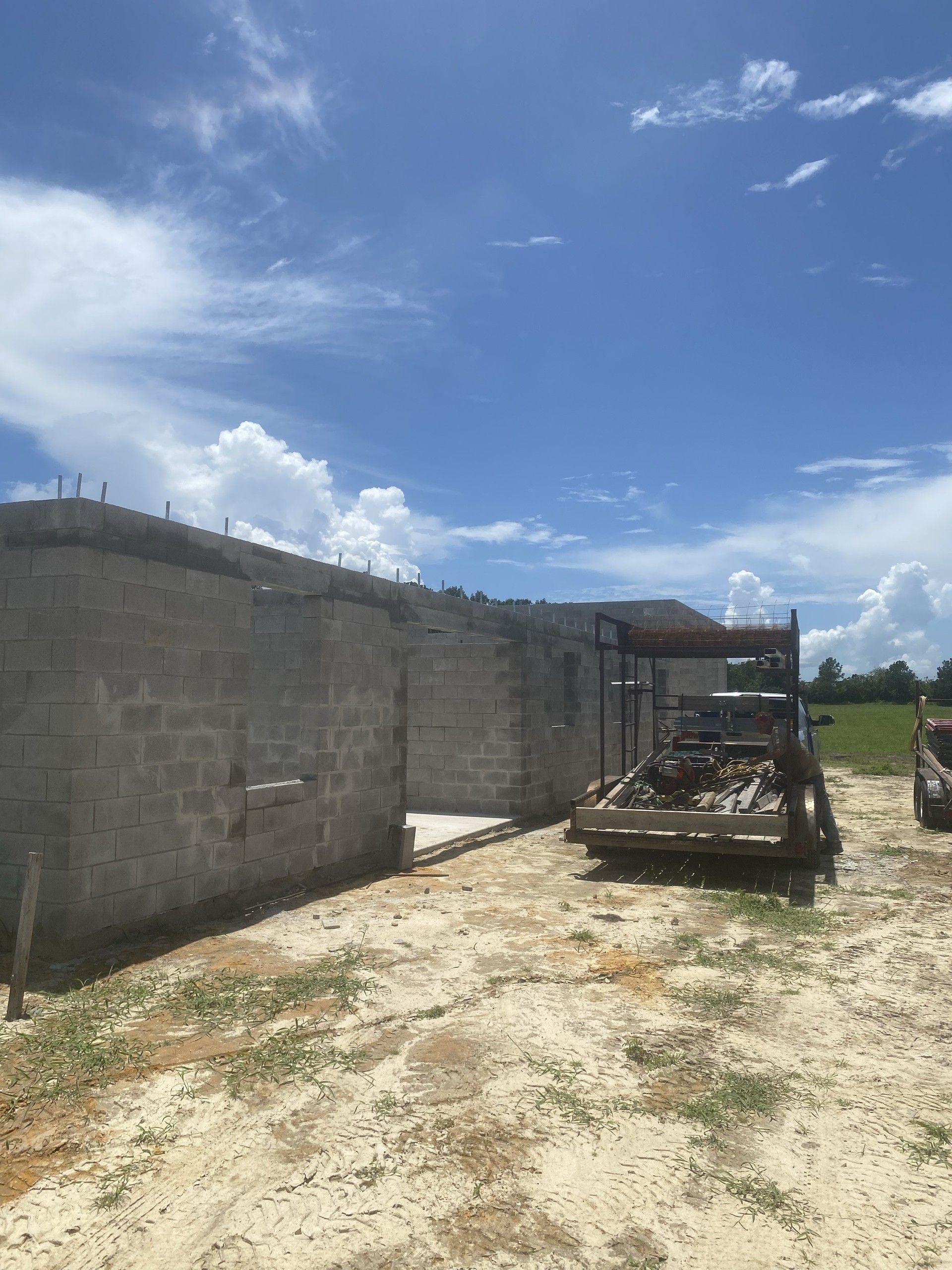 Concrete block walls of a building shell with a loaded equipment trailer and a white van parked on the sand alongside, under a partly cloudy sky
