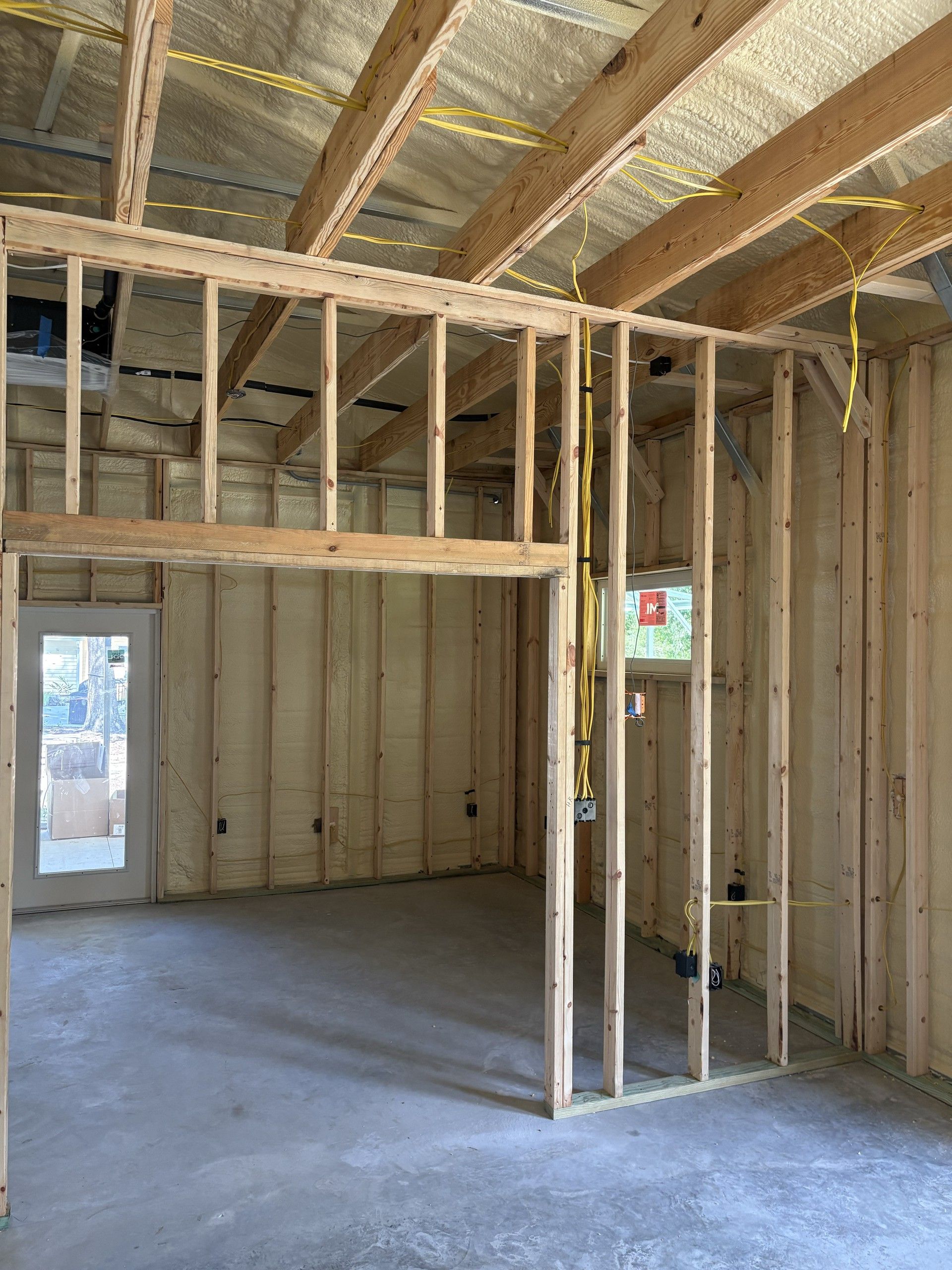 An open room under construction with wood stud walls, a long framed header spanning the space, spray-foam insulated ceiling and walls, a glass exterior door letting in daylight, and a smooth concrete floor