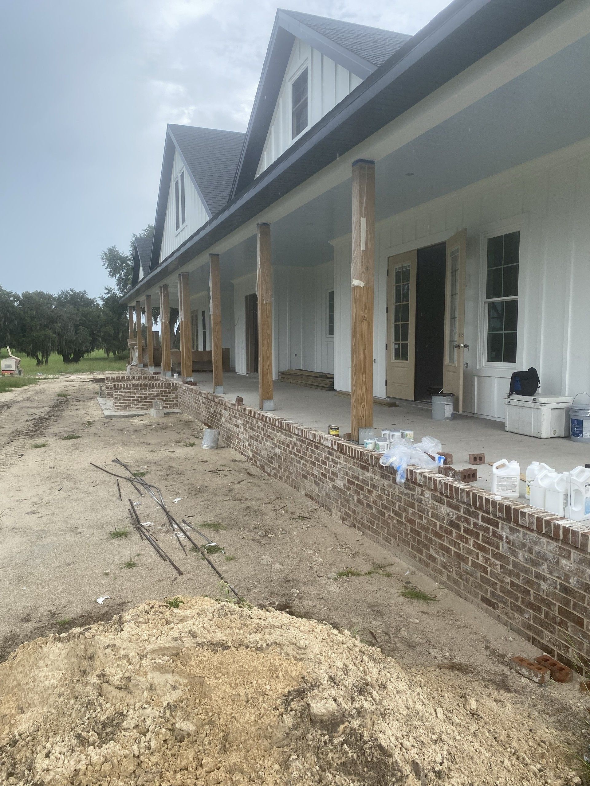 Long view down the covered porch of a white farmhouse under construction, with gabled dormers, wood columns and a brick knee wall running along the edge
