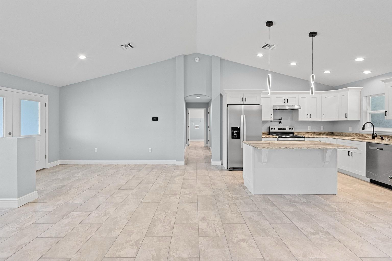Empty room with vaulted ceiling, two pendant lights over a granite-topped kitchen island, stainless refrigerator, and an arched hallway opening