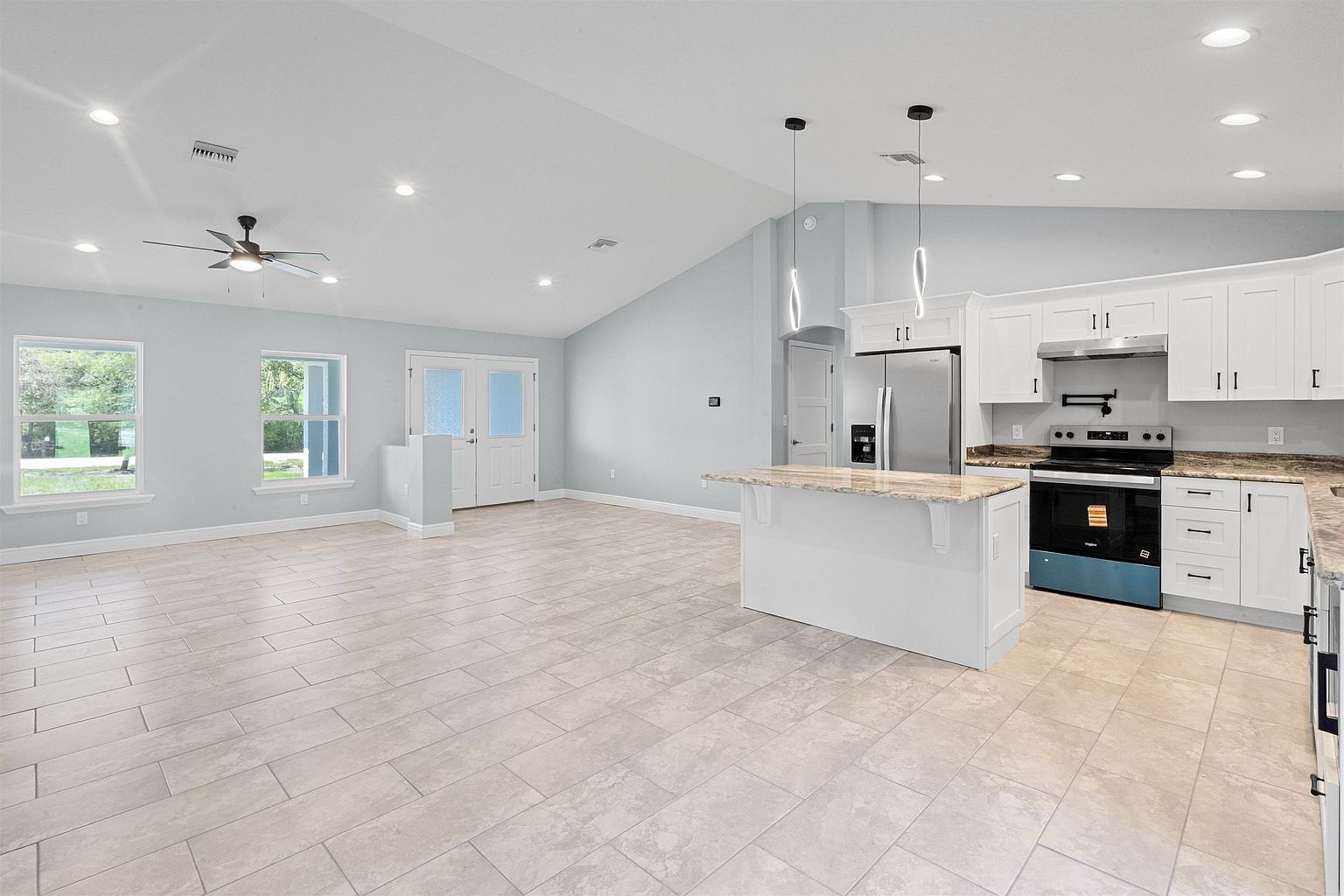 Empty living space with ceiling fan and two windows on the left, adjoining a white kitchen with island, range and refrigerator on the right