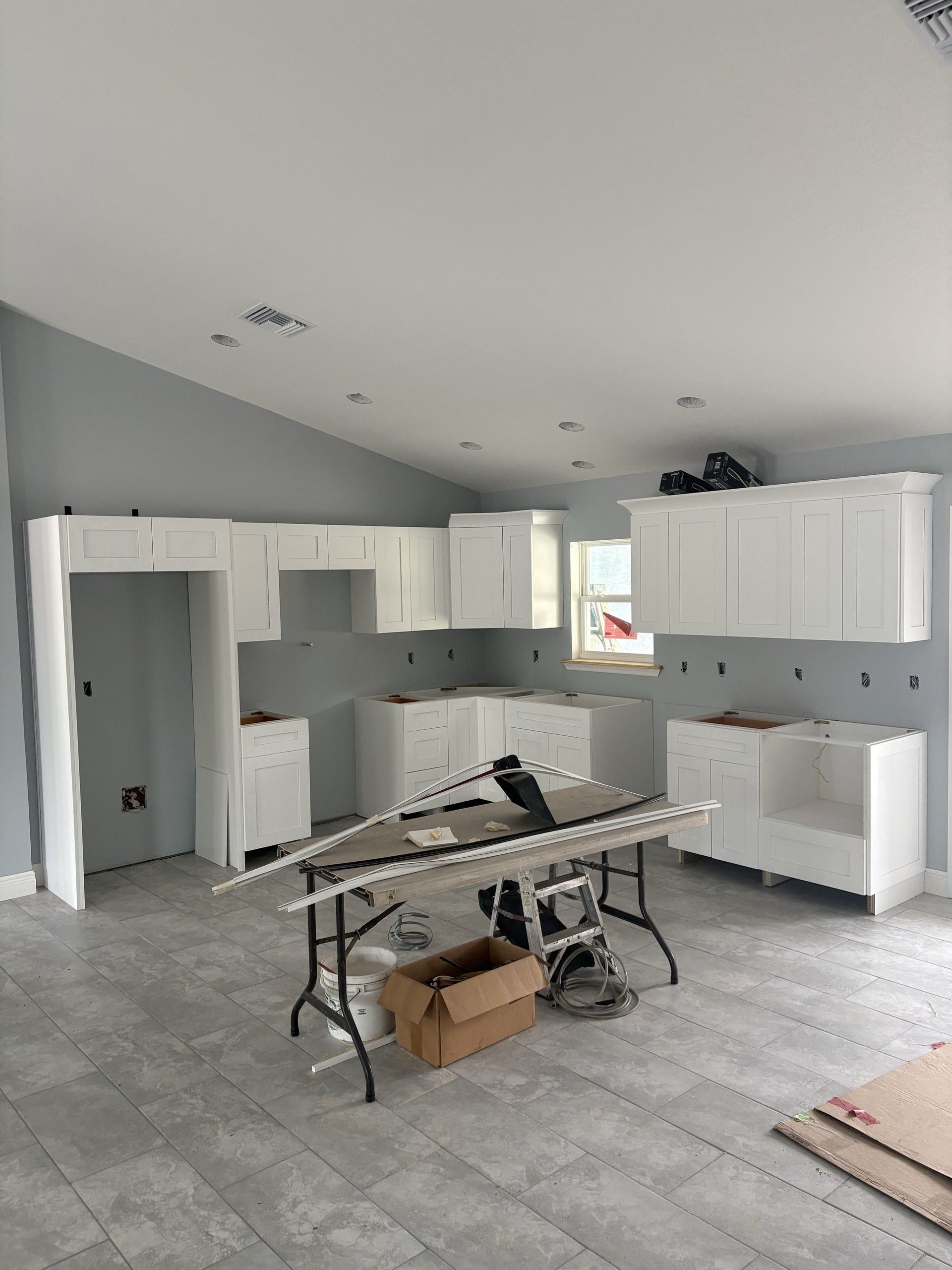 Wide view of a kitchen with white shaker upper and base cabinets, gray tile floor, and a folding table holding trim pieces
