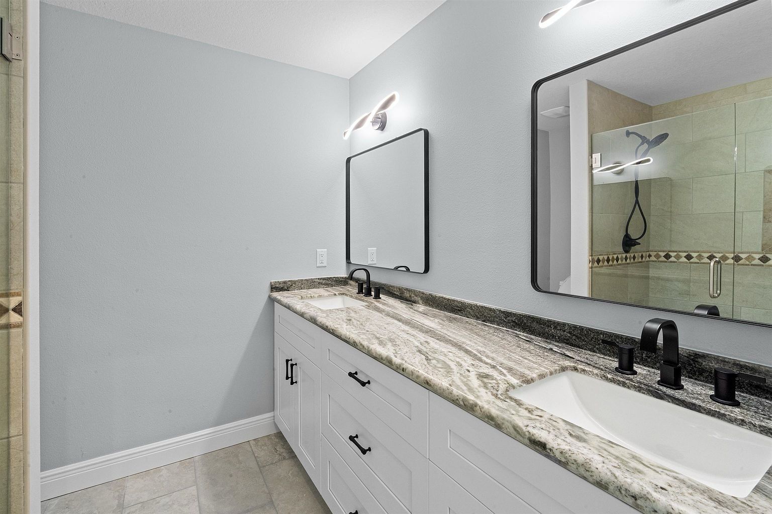 Primary bathroom with a long granite double vanity, white drawers, black faucets, two black-framed mirrors and wall sconces; a tiled shower is reflected in the mirror