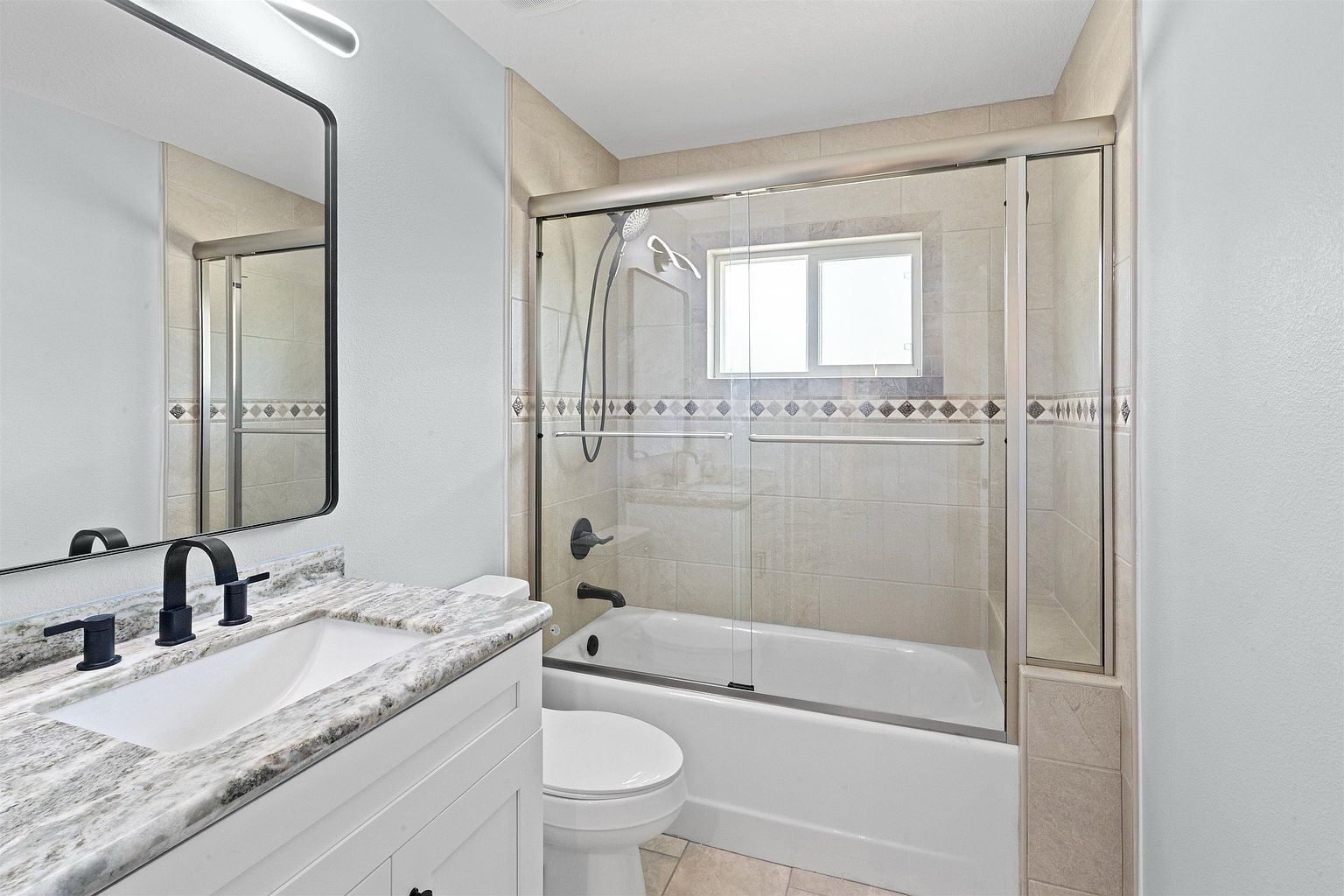 Bathroom with granite-topped white vanity, black faucet, framed mirror, toilet, and a tiled tub-shower with sliding glass doors and a decorative tile border
