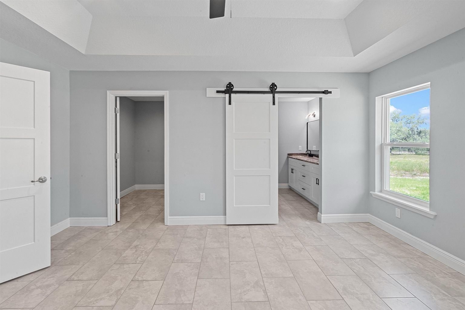 Empty primary bedroom with tray ceiling, a doorway to a walk-in closet, a sliding barn door revealing a bathroom vanity, and a window looking onto open grassland