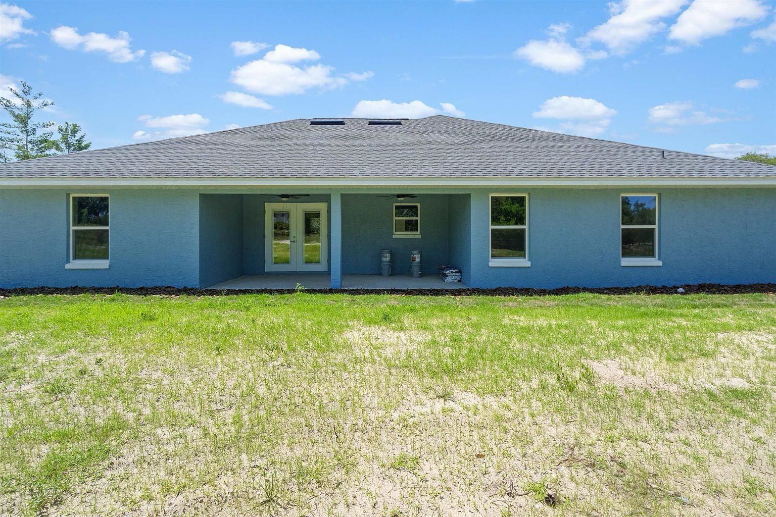Straight-on rear view of a light blue stucco house with a wide hip roof, recessed covered patio with French doors and ceiling fans, and two paint buckets on the slab