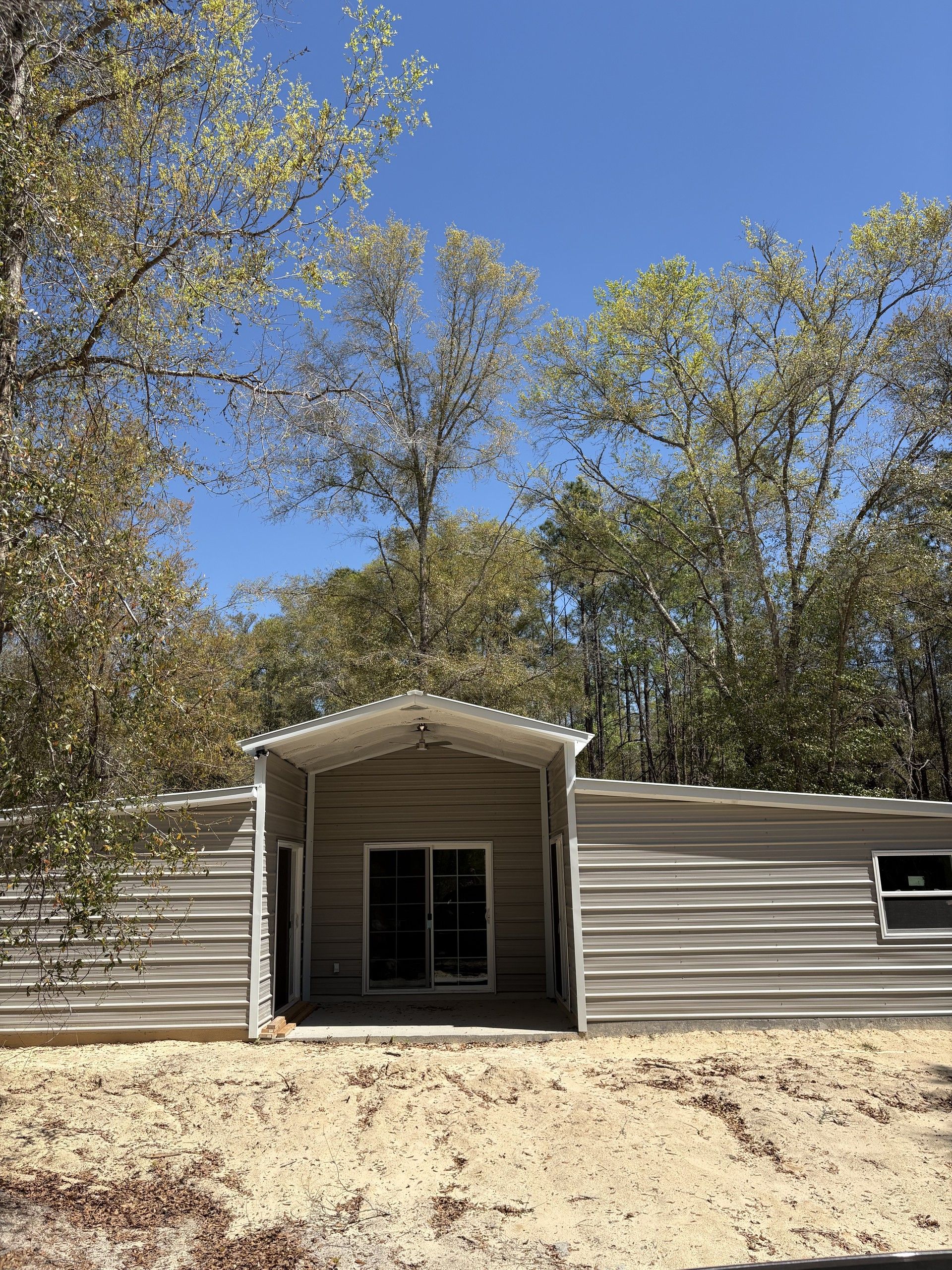A completed tan metal barndominium with a gabled recessed porch at center, ceiling fan under the porch, a sliding glass door and flanking entry doors, wings extending to both sides, sandy ground in front and tall trees behind under a blue sky