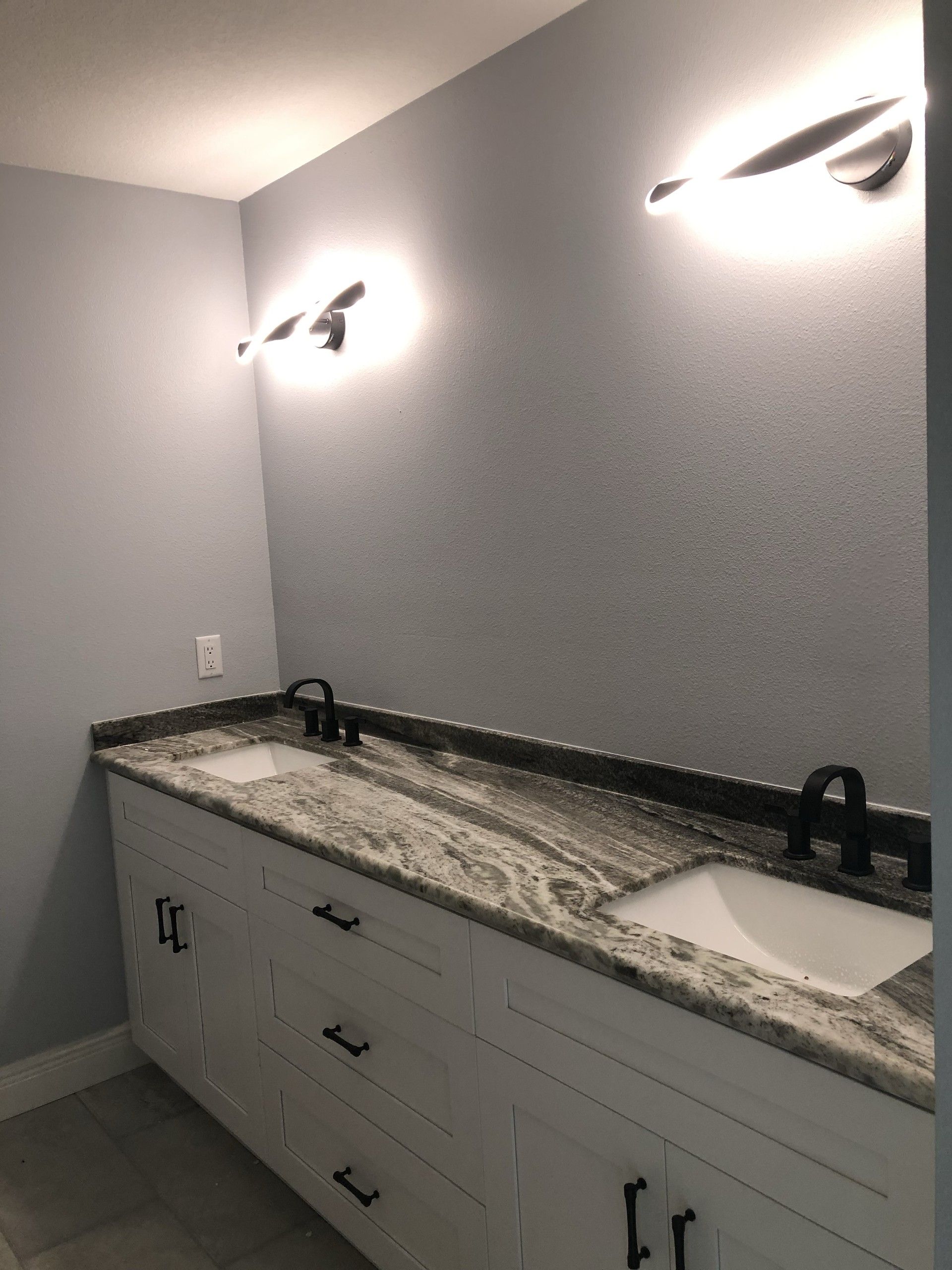 Bathroom double vanity with white cabinets, a granite countertop, two undermount sinks, black faucets, and two lit wall sconces