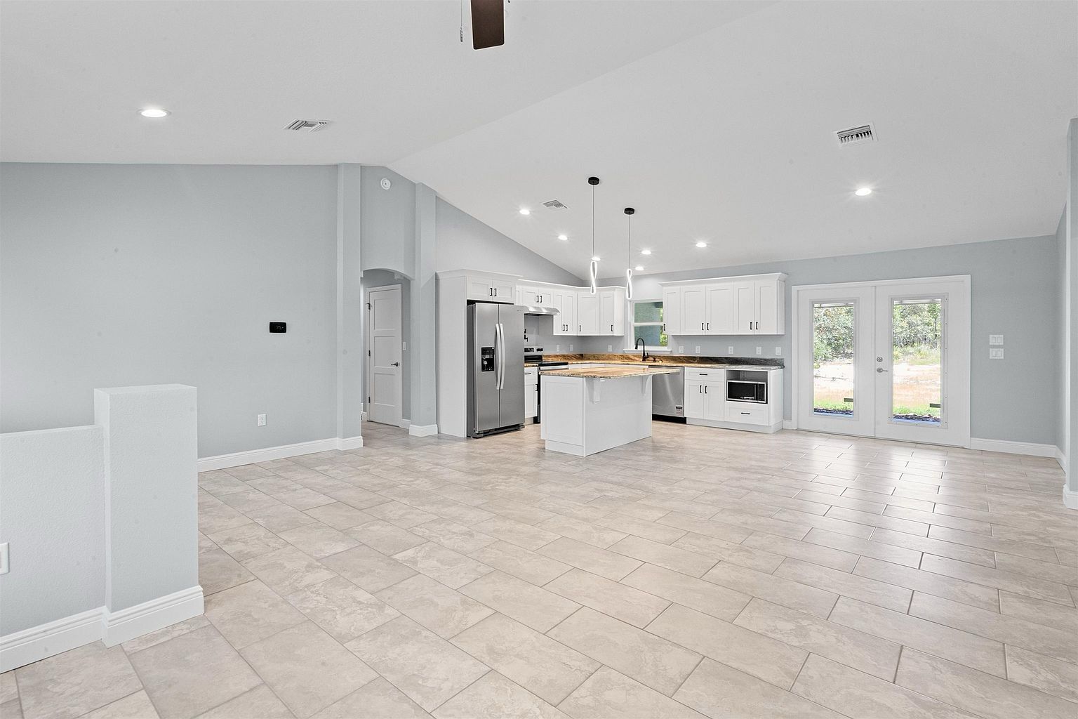 Vaulted empty living room with ceiling fan, recessed lights, and a white kitchen with island and French doors along the far wall