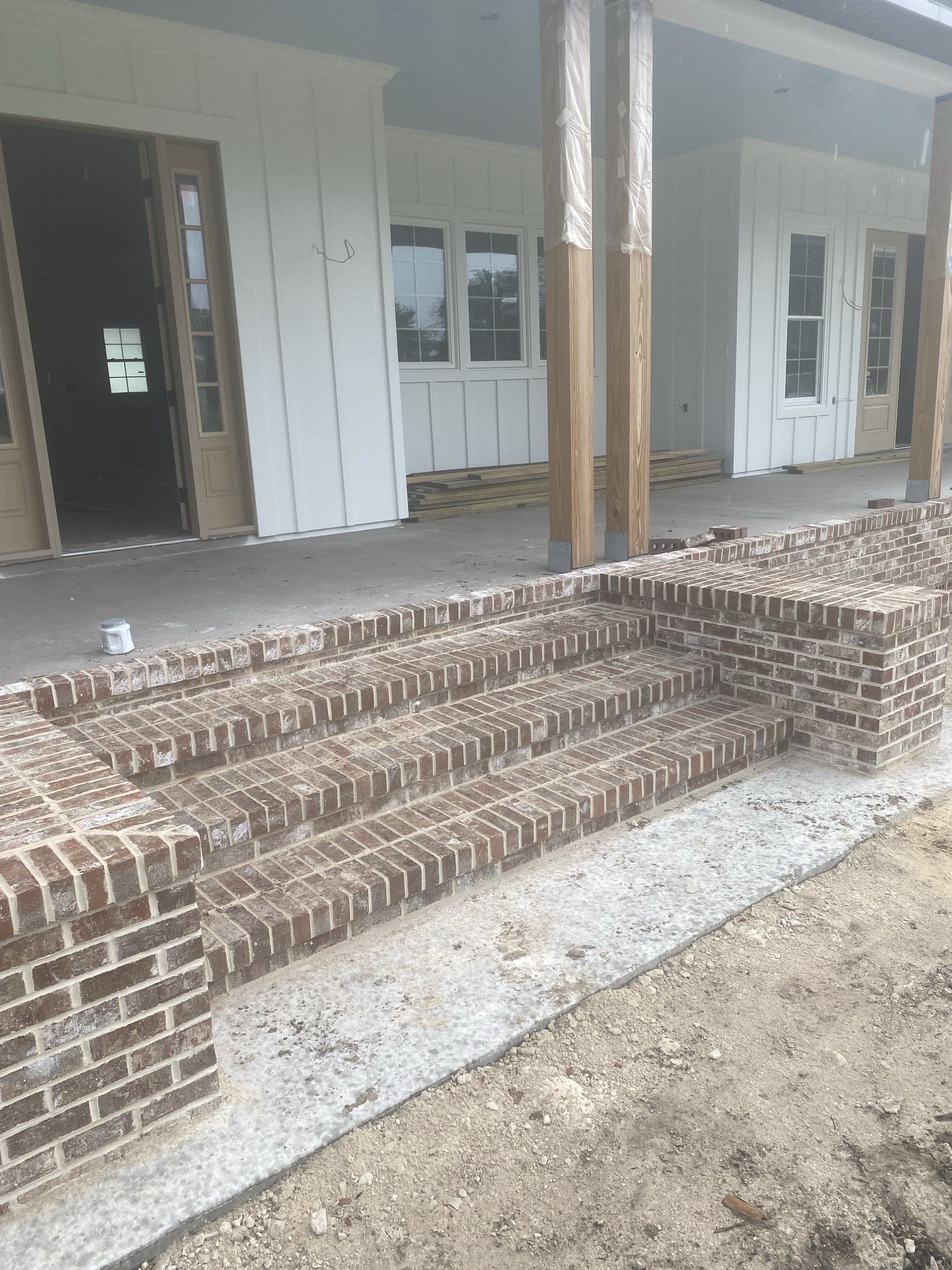 Brick front steps and a low brick wall leading up to a covered porch on a white board-and-batten house under construction, with plastic-wrapped wood porch columns