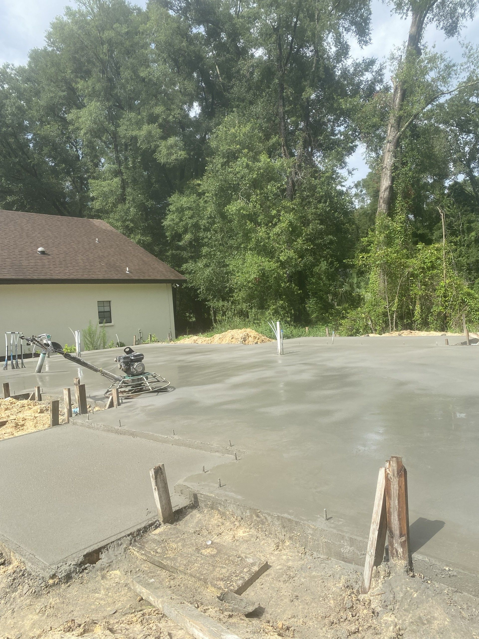A freshly troweled concrete slab poured next to an existing single-story stucco house, with a ride-behind power trowel parked on the wet surface and woods behind