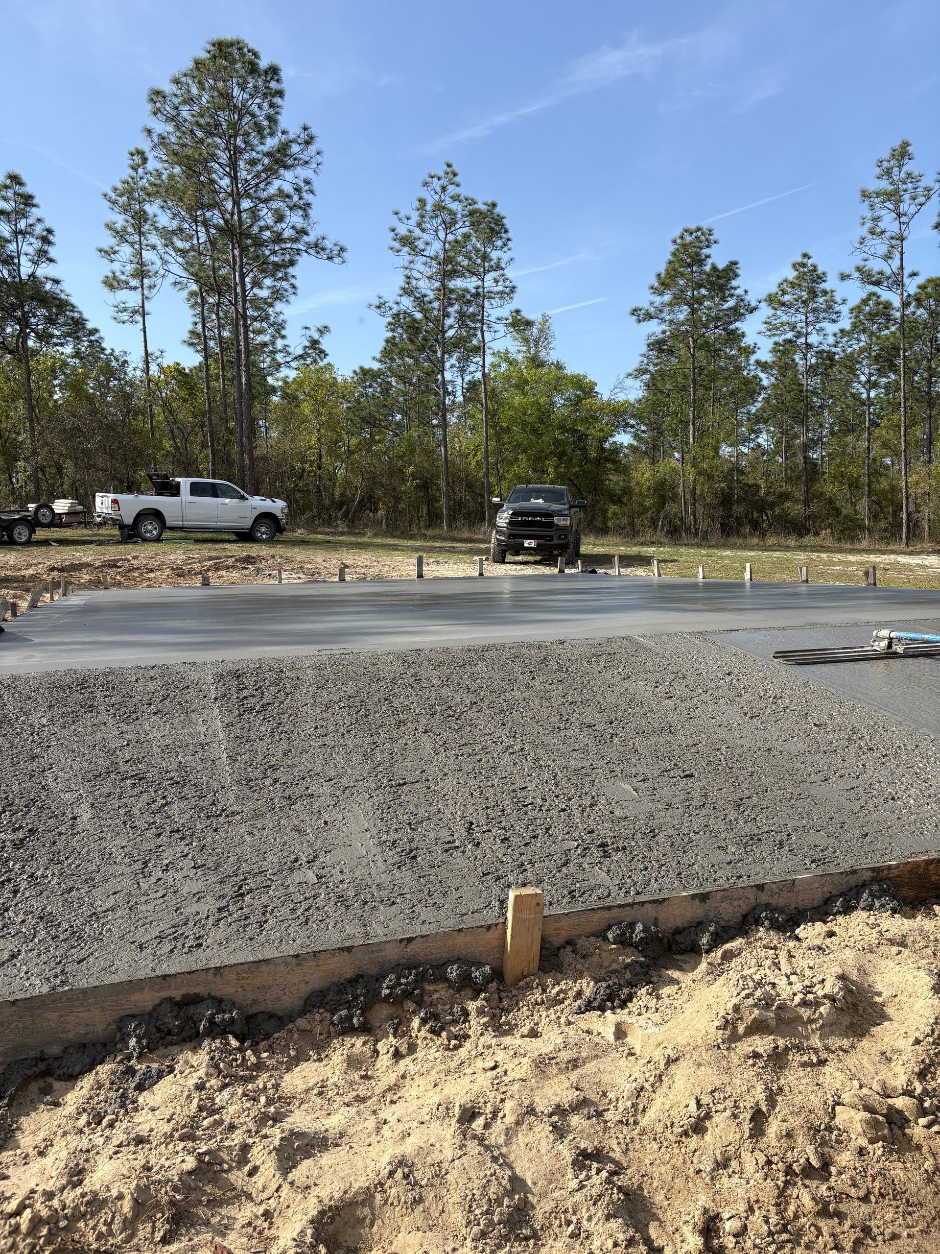 A freshly finished concrete slab with a rough exposed edge and wood form boards, with two pickup trucks, a utility trailer and pine trees under a blue sky behind it