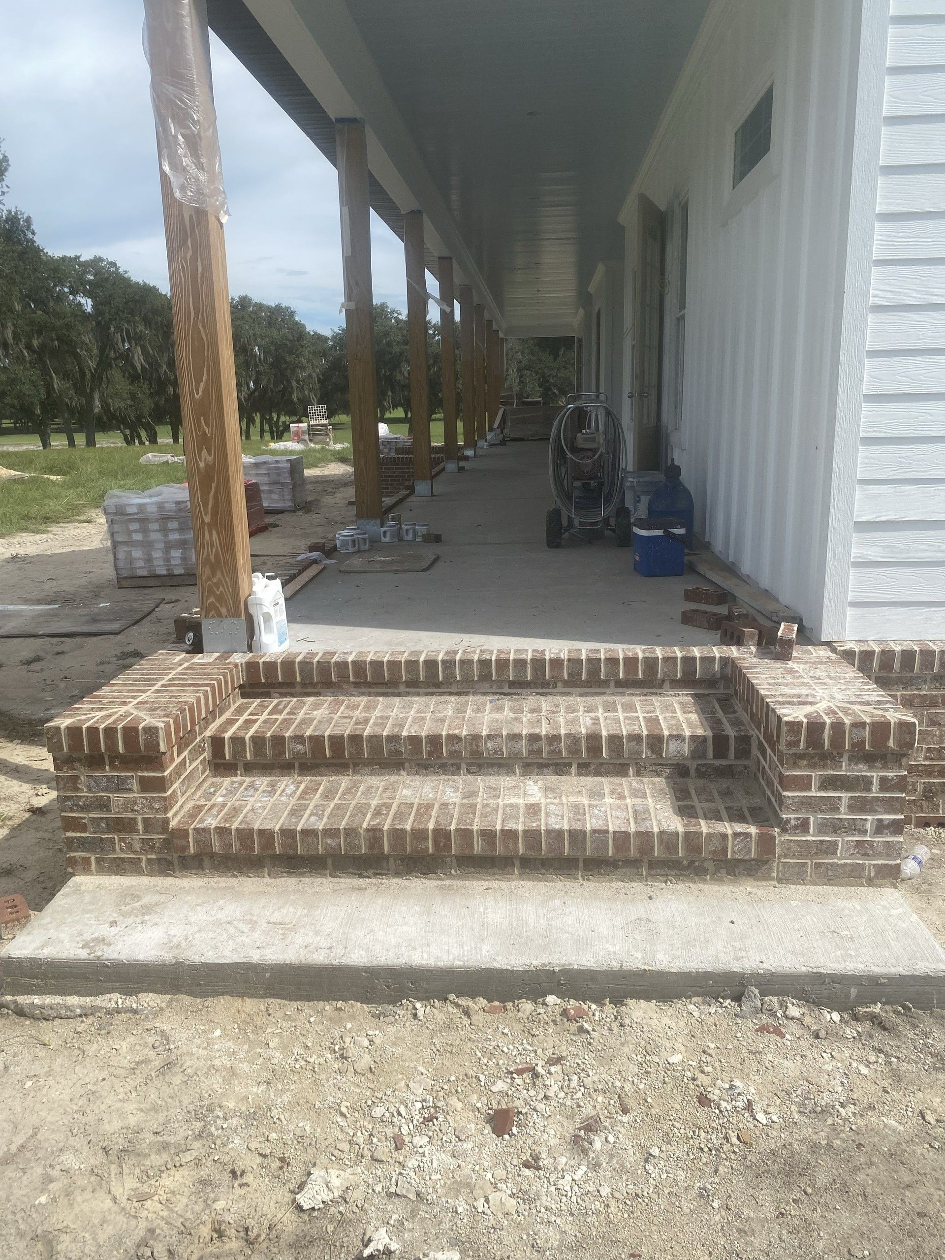 Brick steps with flanking brick cheek walls leading up to a concrete porch, with live oak trees visible beyond the porch columns