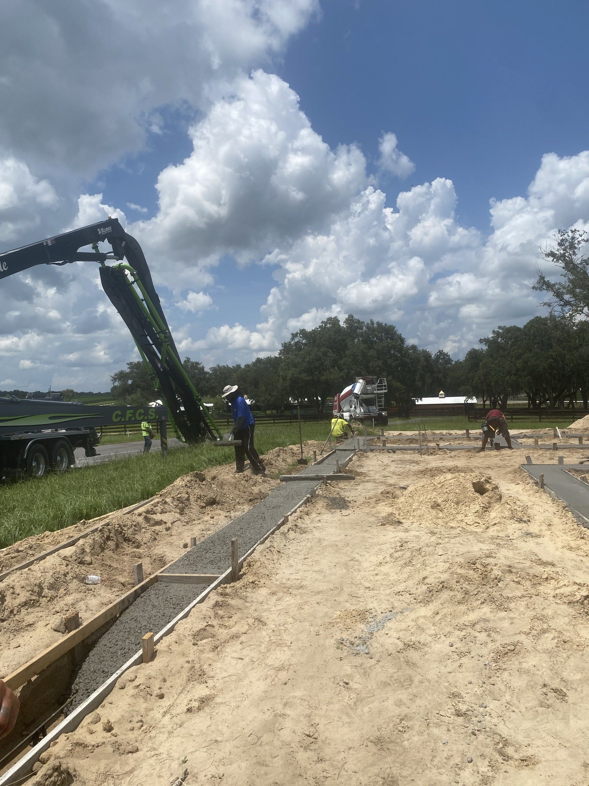 A worker in a wide-brimmed hat pulls a screed along a long footing form filled with wet concrete, with a pump truck, a mixer truck and other crew members behind