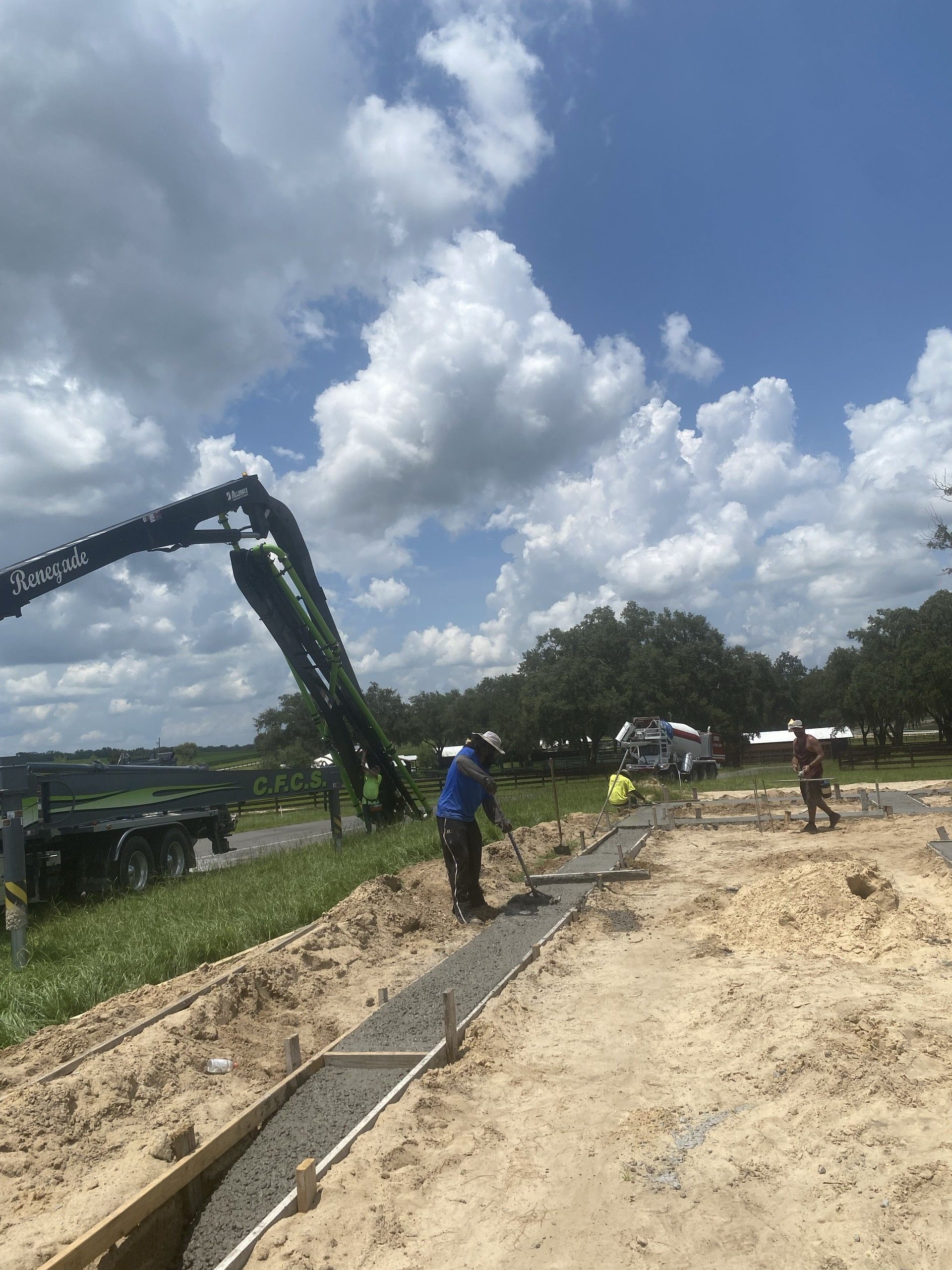 A worker in a blue shirt and sun hat rakes wet concrete along a footing form, with a concrete pump truck, a mixer truck and other crew visible under a cloud-filled sky