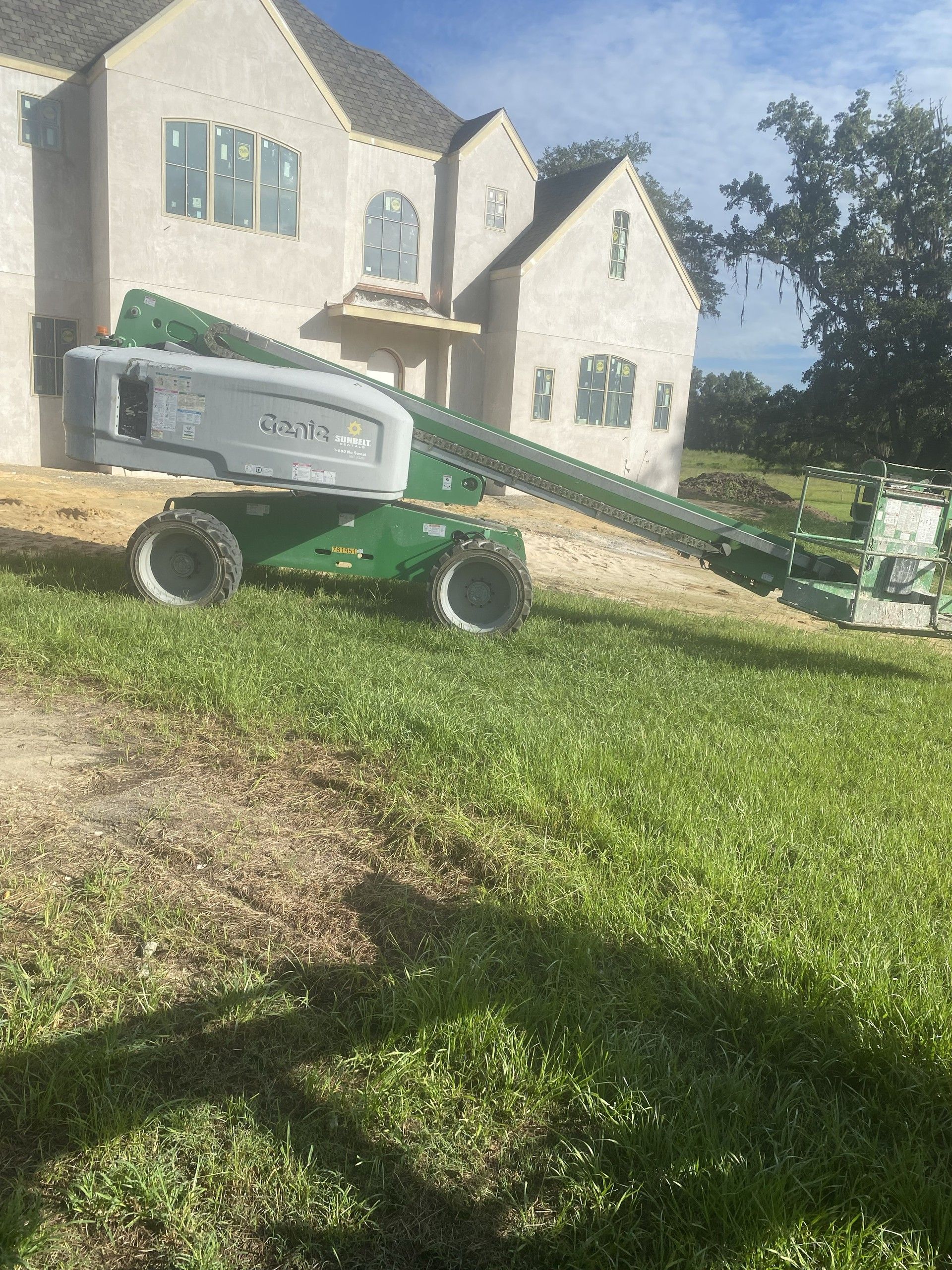 A large two-story stucco custom home with steep gables and arched windows nears completion, with a green articulating boom lift parked on the grass in front of it