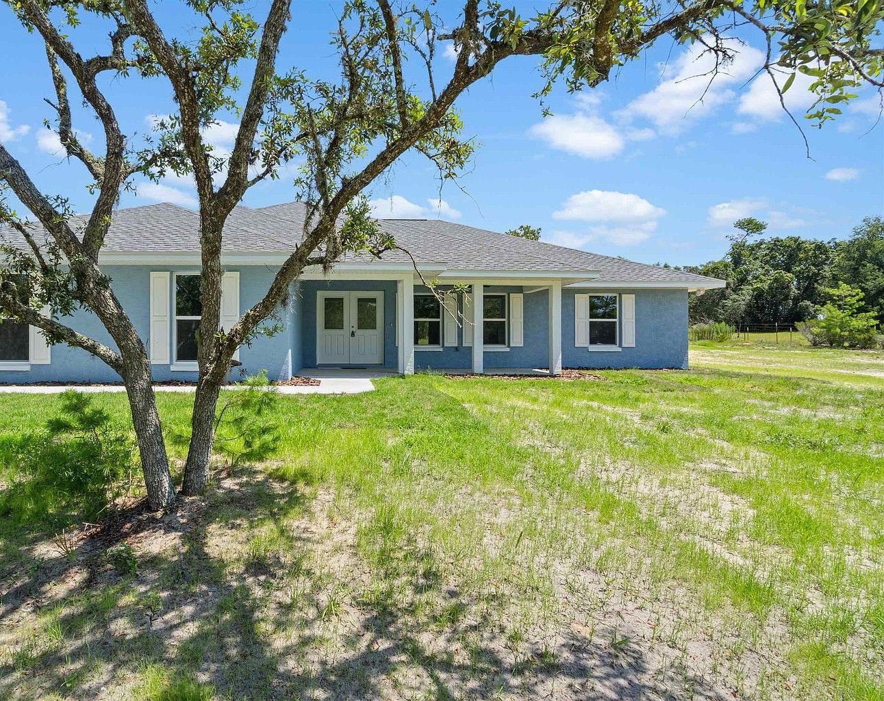 Light blue stucco single-story house with white columns and covered entry porch, seen through the spreading branches of a large live oak in the front yard