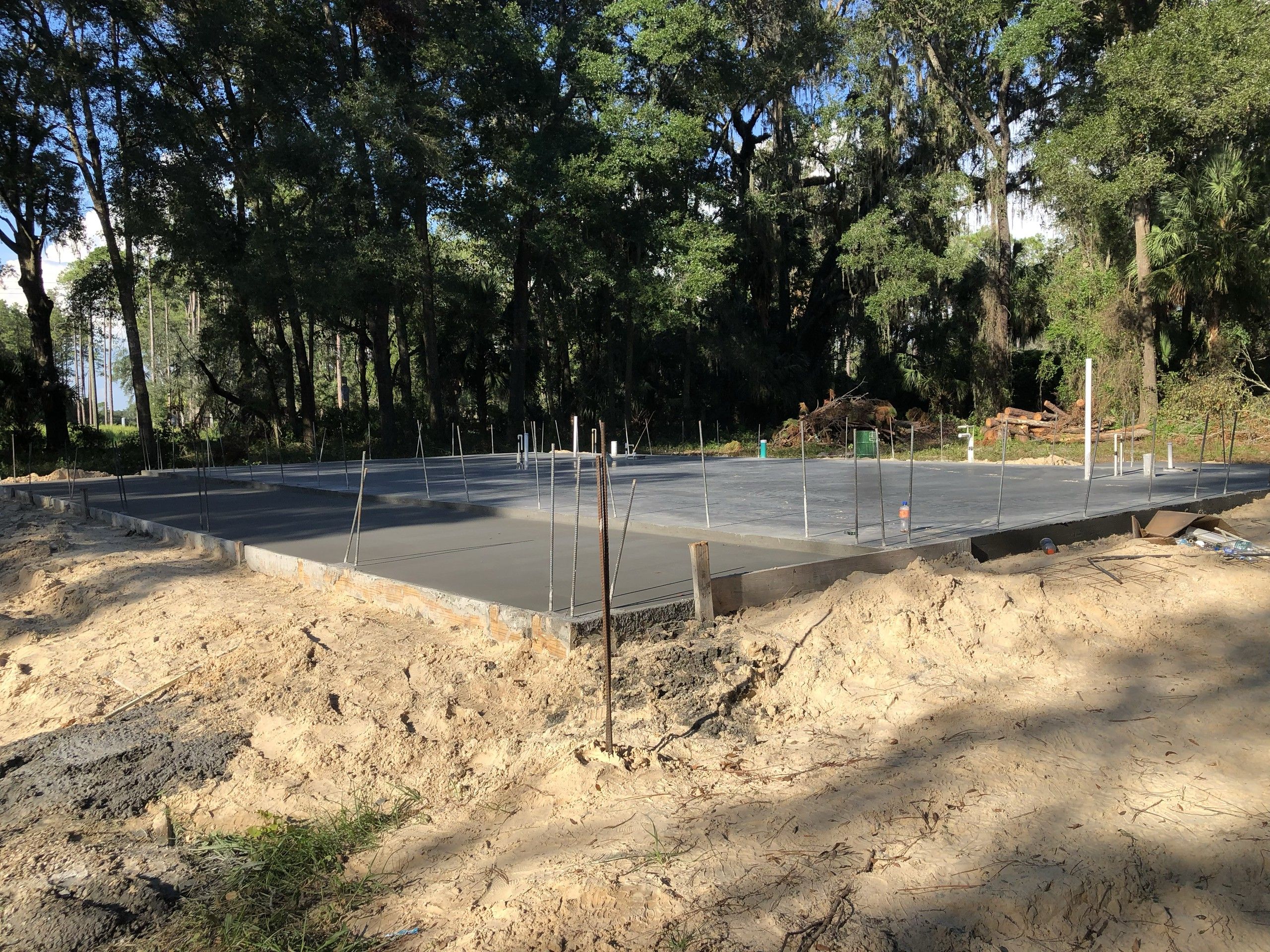 A large finished concrete slab with a formed stem wall and rebar dowels sits on a cleared lot ringed by oak and palm trees, with cleared brush piled at the tree line