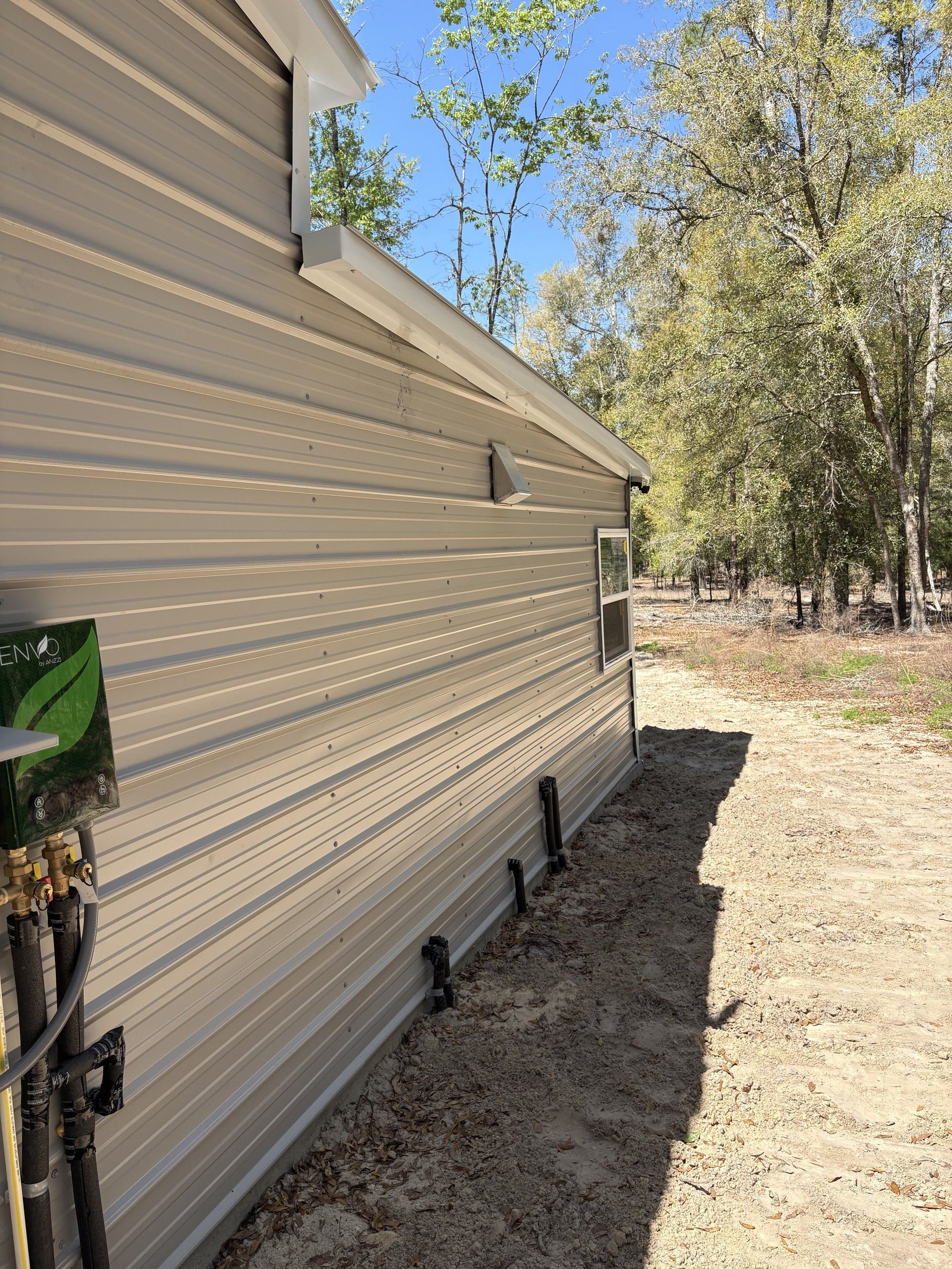 The long side wall of a tan metal building with white gutters and a gable end, plumbing stub-outs along the base, sandy soil in front and oak and pine woods under a clear blue sky