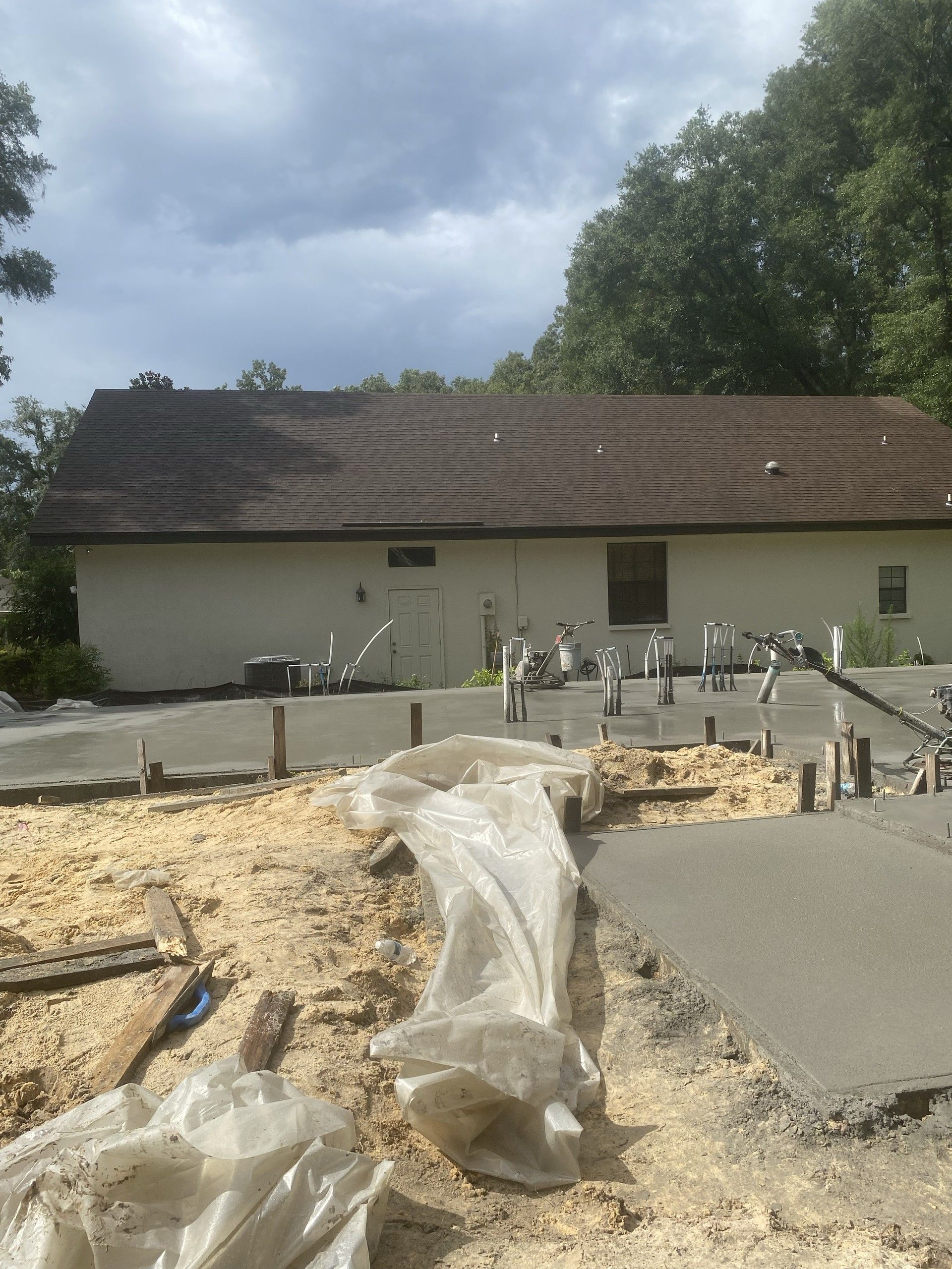 The rear elevation of a single-story stucco house with a freshly poured concrete slab in front of it, plumbing stub-ups rising through the concrete and plastic sheeting on the sand