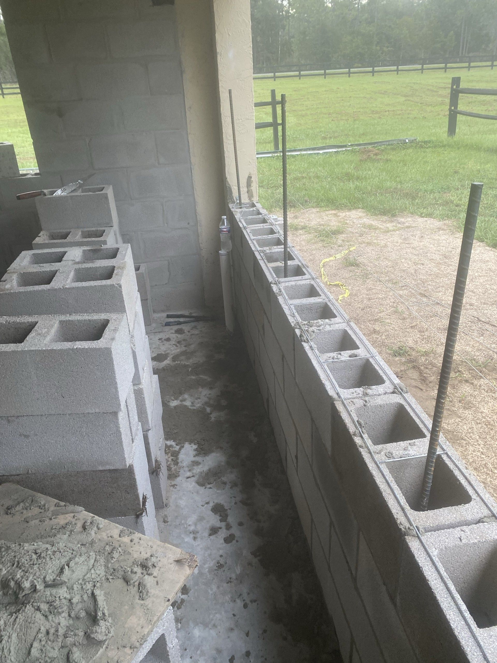 A course of concrete block laid with rebar and wire reinforcement under a covered porch, with stacked block, a trowel and a mortar board nearby and pasture beyond