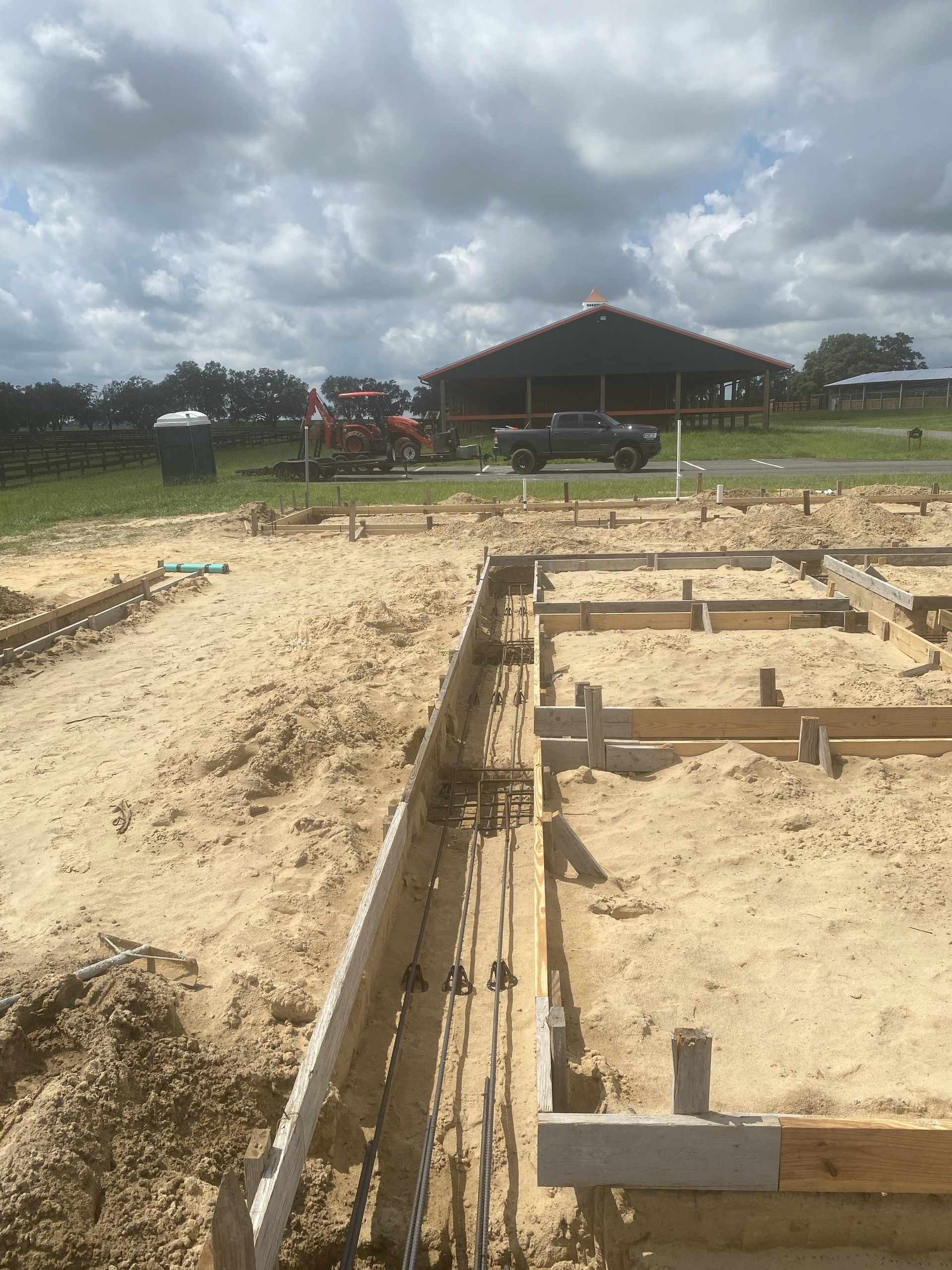 Wooden footing forms filled with sand run across the frame with rebar cages and tensioning cables laid in the trenches, and a large open equestrian pavilion in the background