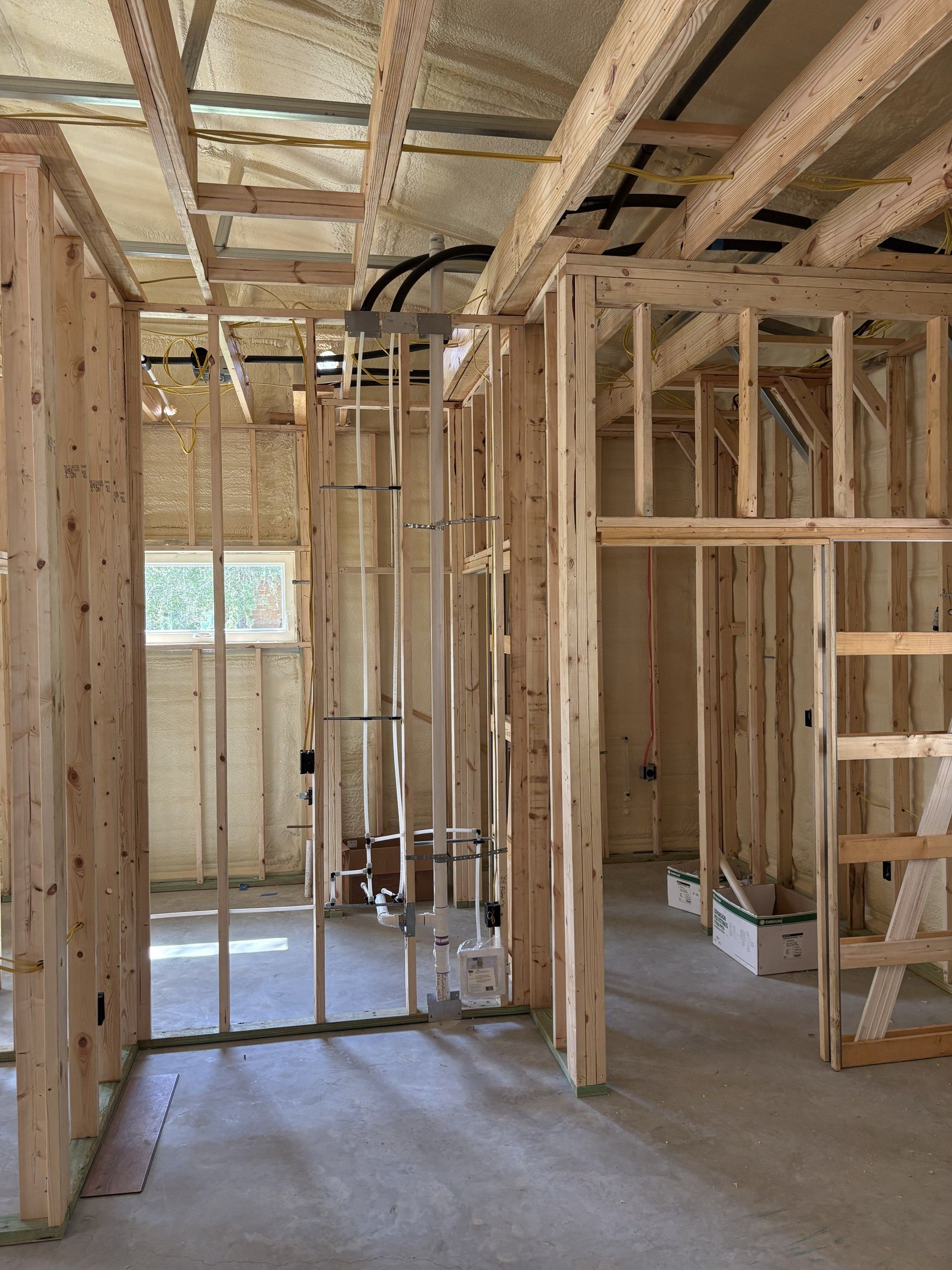 Interior framing of a bathroom under construction, with white PVC drain and vent pipes rising between wood studs, insulated walls and ceiling, a small window, and cardboard supply boxes on the concrete floor