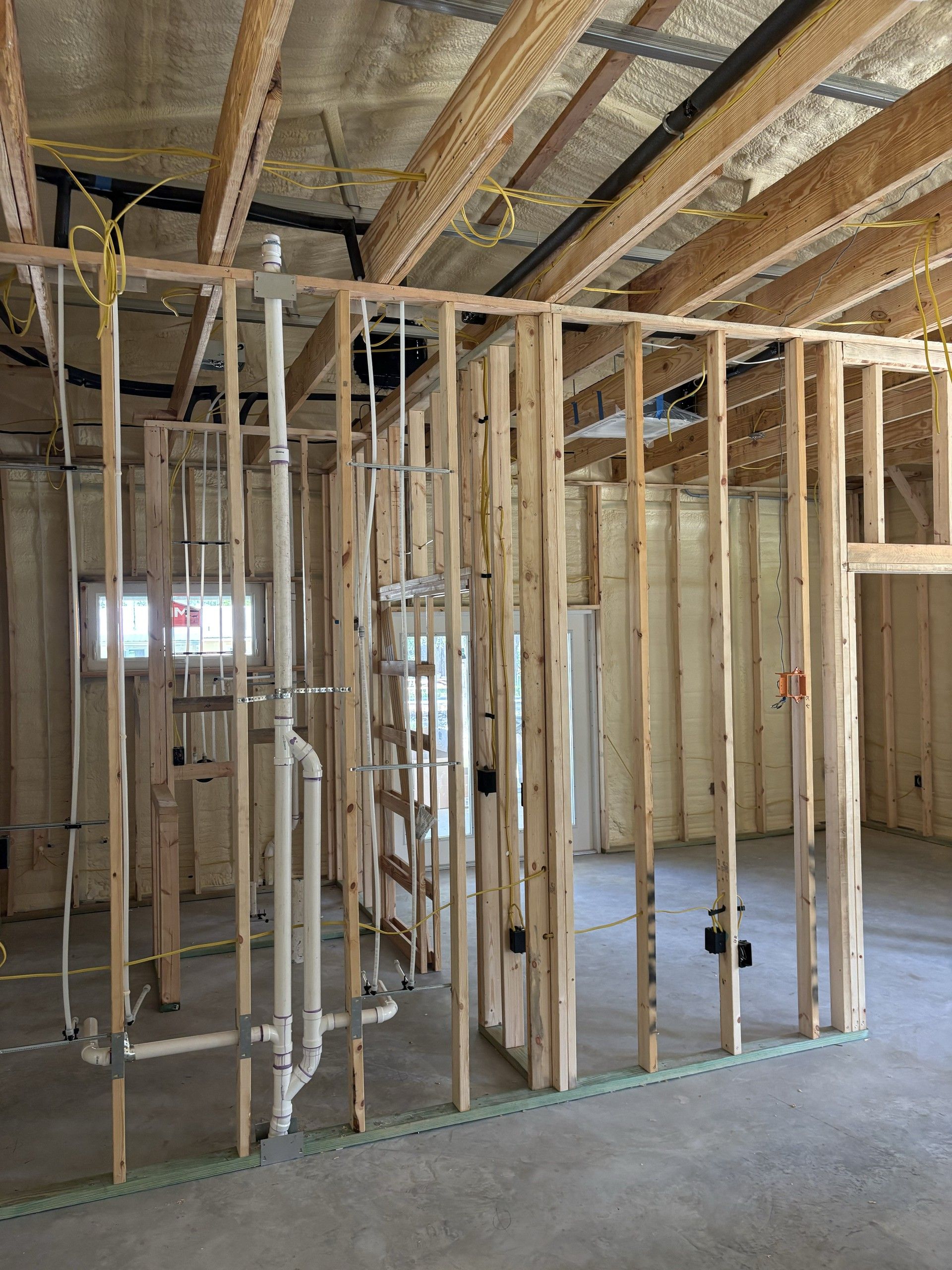 Wood-framed interior walls with a tall white PVC vent stack and white PEX water lines running vertically between studs. Insulated ceiling, exposed beams, and a concrete floor fill the rest of the frame