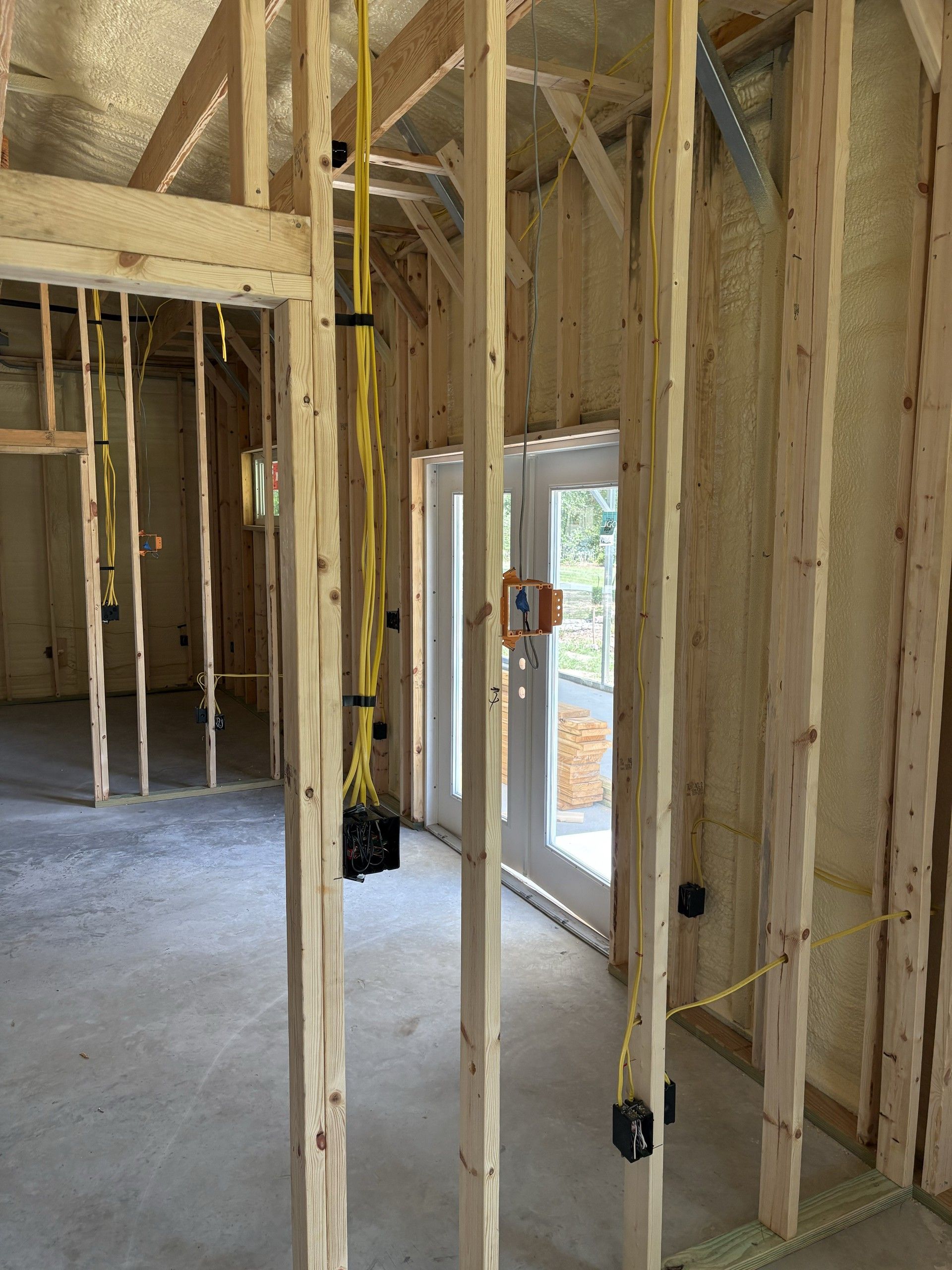View through framed wood stud walls toward a glass exterior door, with bundles of yellow electrical cable running down a stud and black outlet boxes mounted at wall height above a concrete floor