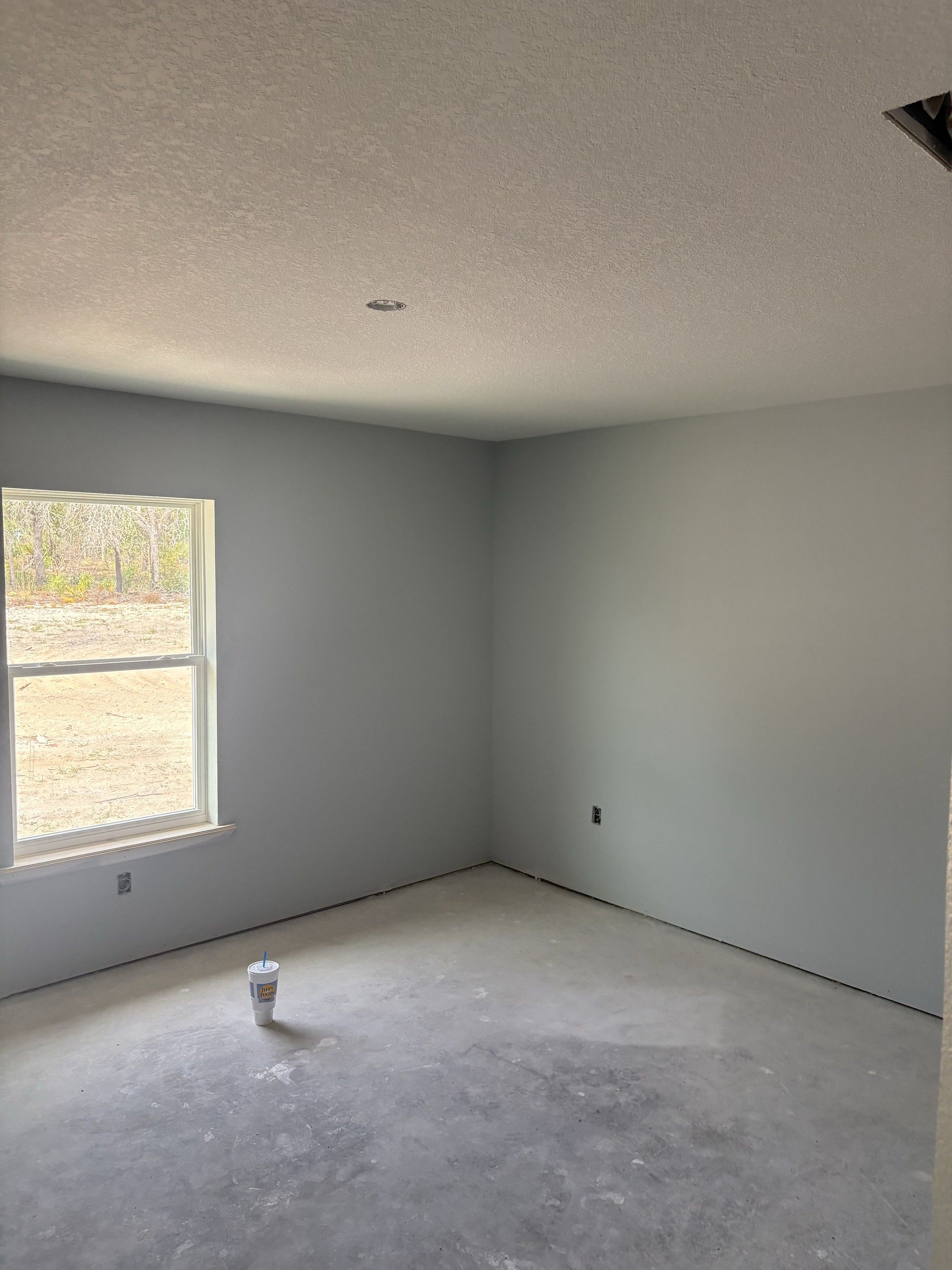 Empty bedroom with light gray painted walls, a double-hung window, textured ceiling, and a bare concrete floor with a paper cup on it