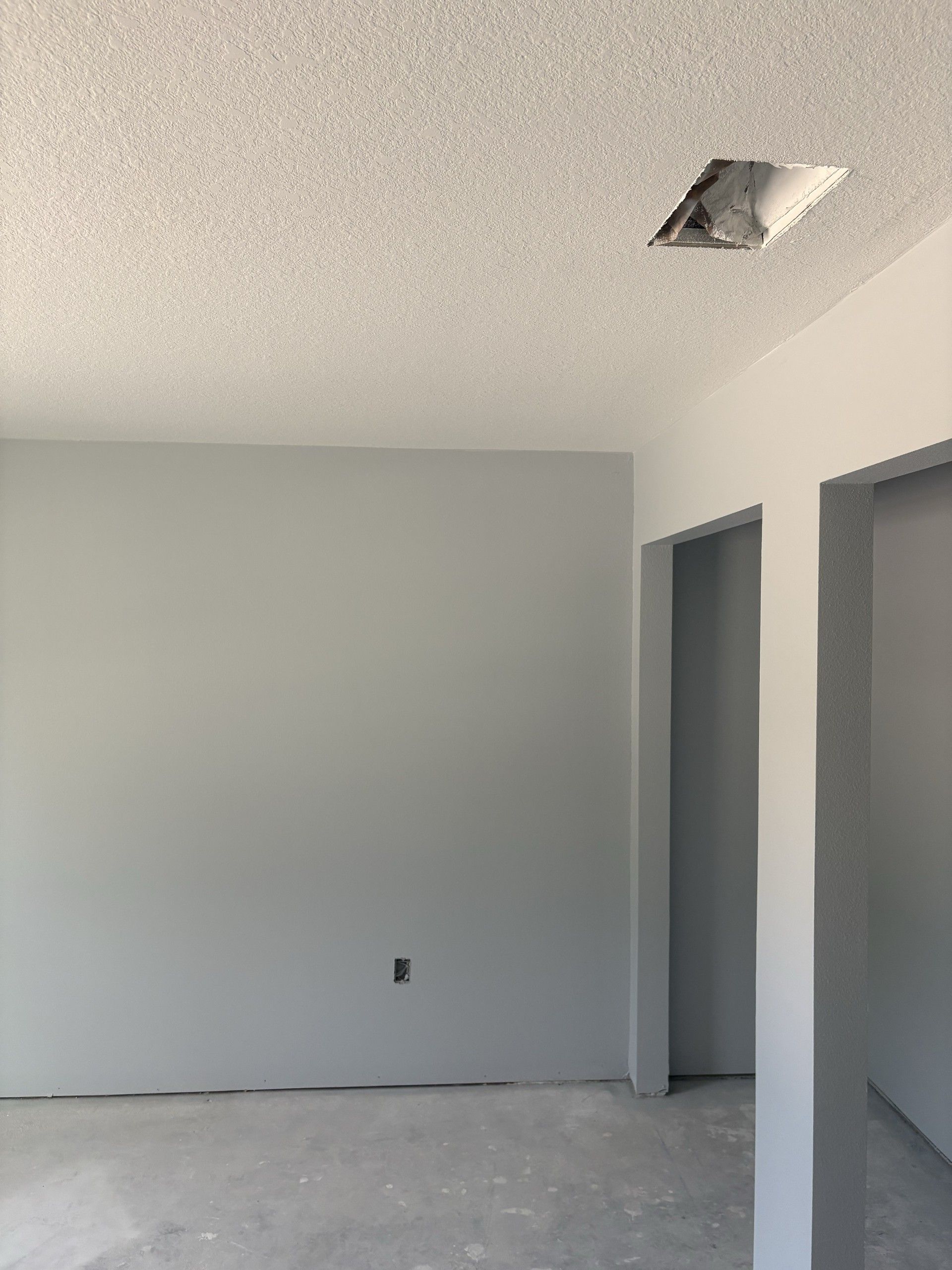 Corner of an unfinished room with painted walls, an open ceiling duct opening, two doorway openings, and a concrete floor