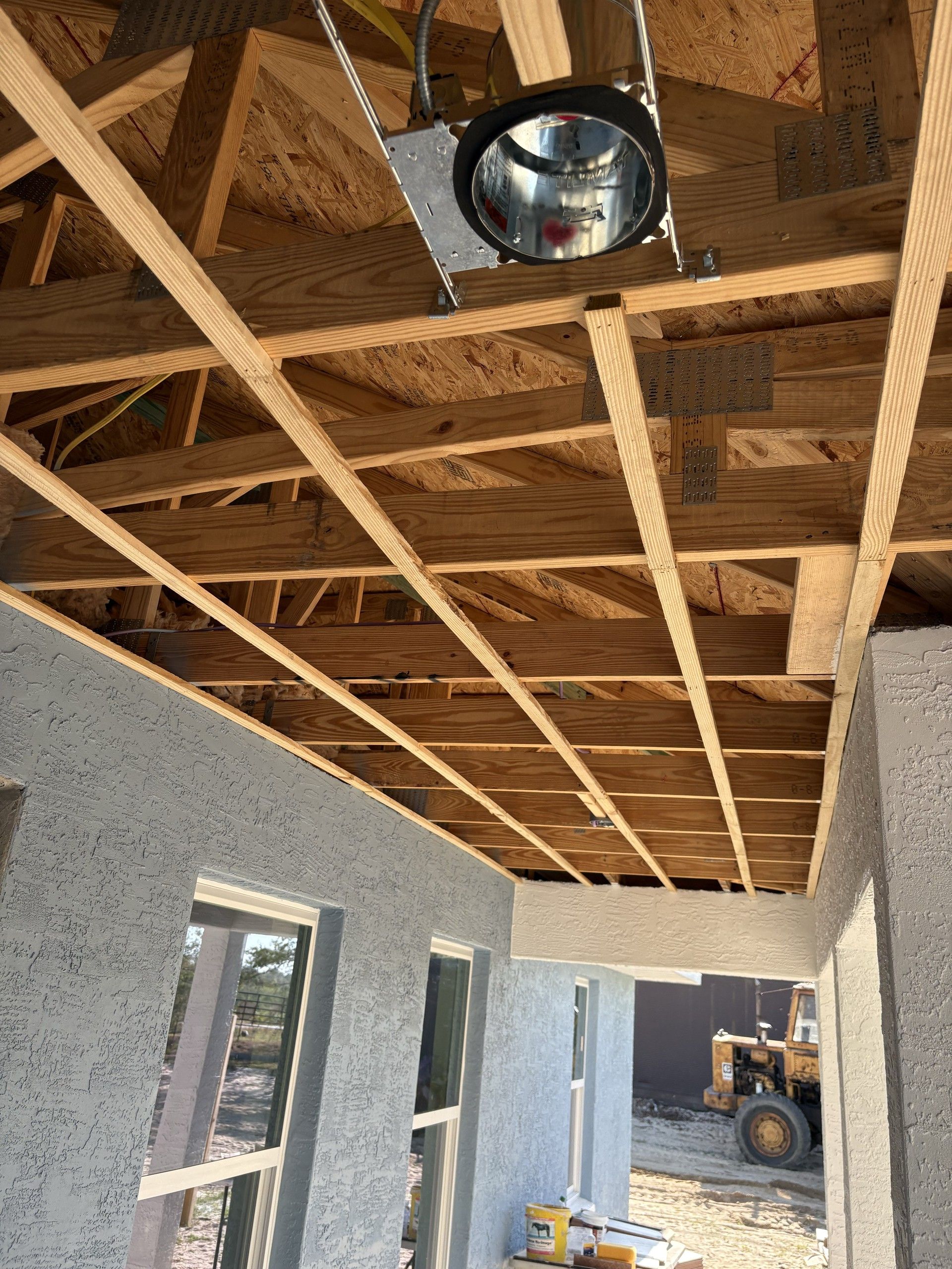 Exposed ceiling joists and roof sheathing over a covered porch, with a recessed light can installed and a tractor parked outside